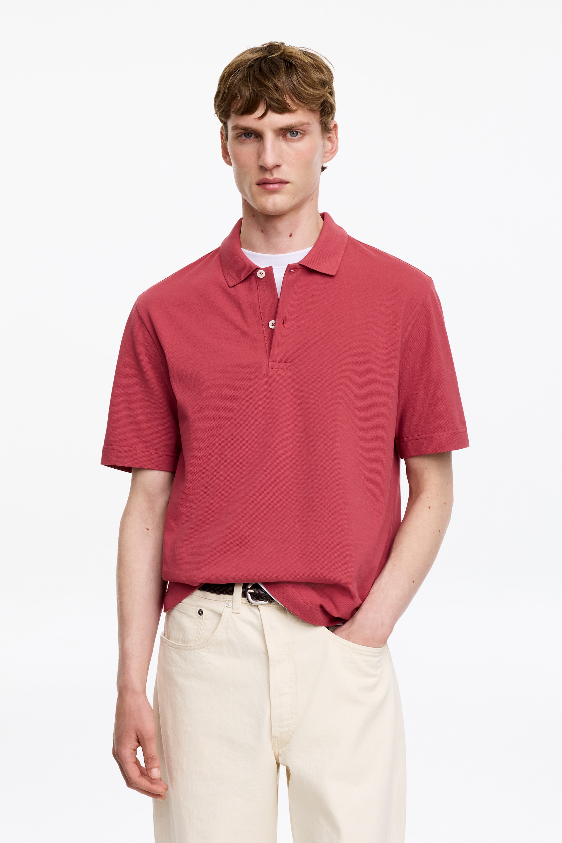 View larger image: A red polo shirt is being worn, facing forward, featuring a classic polo collar, a two-button placket with white buttons, and short sleeves. It is layered over a white crew neck t-shirt.