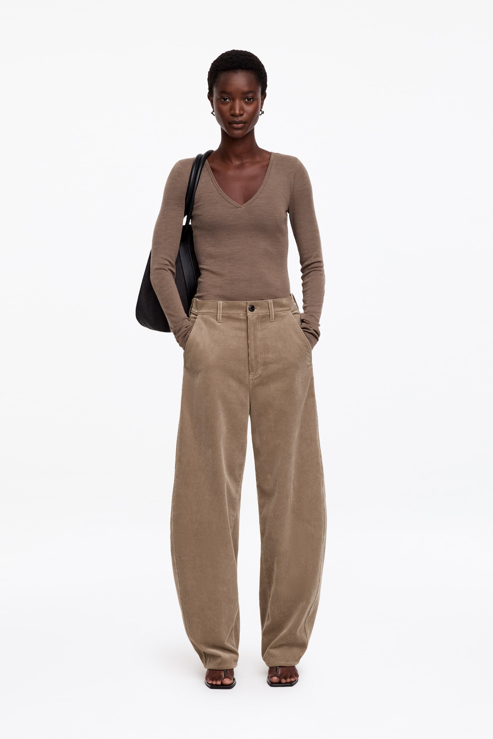 Agrandir l'image: Pantalon en velours côtelé marron clair à silhouette barrel leg, avec poches avant et taille normale, porté par le mannequin qui se tient debout, les mains dans les poches. Le pantalon est associé à un haut marron ajusté à manches longues et col V, ainsi qu’à un sac à bandoulière noir.