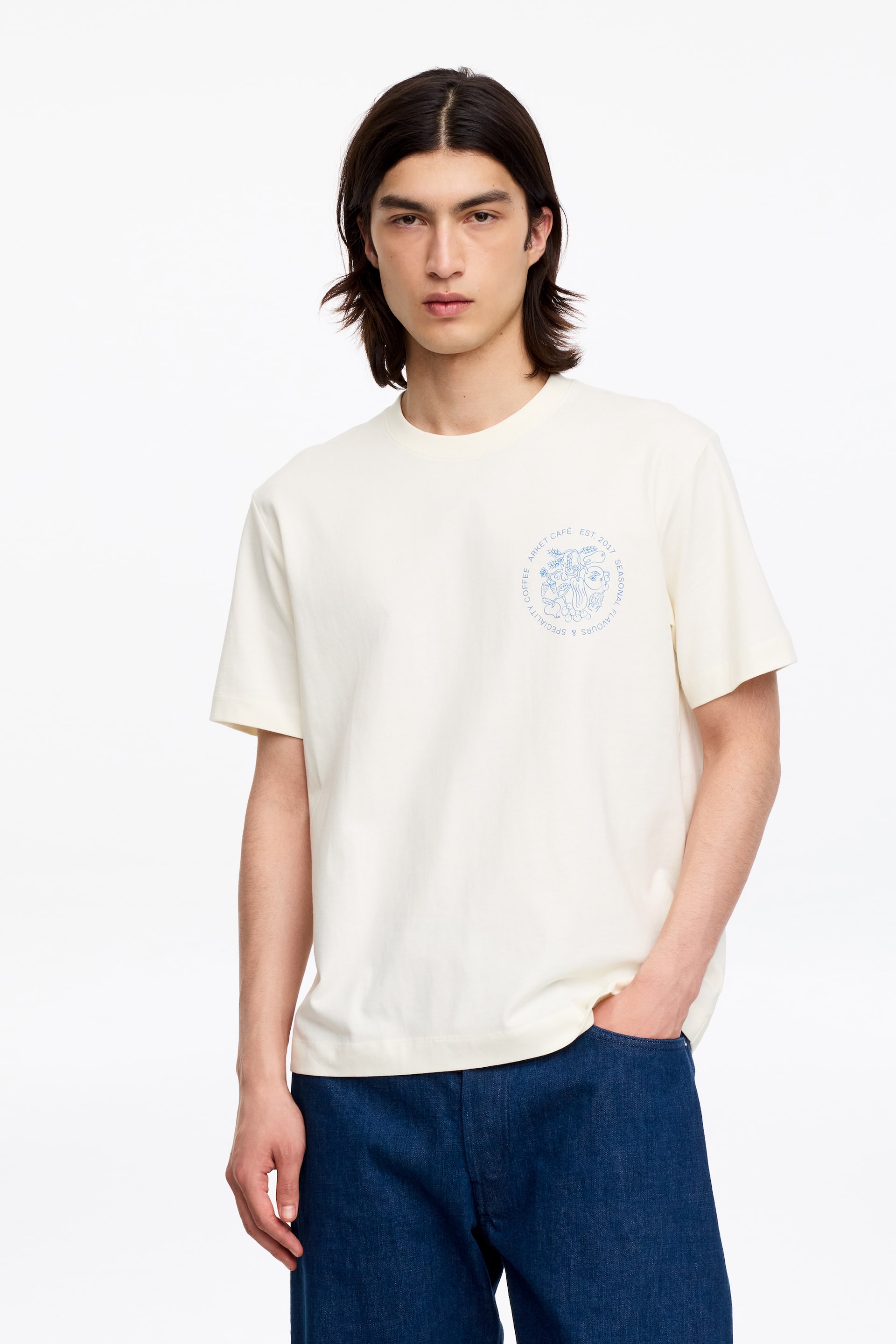 Grotere afbeelding bekijken: Een crèmekleurig t-shirt met een lichtblauw cirkelvormig grafisch ontwerp op de linkerborst, met tekst en gestileerde elementen. Het is ontworpen met een ronde hals en korte mouwen, naar voren gericht.