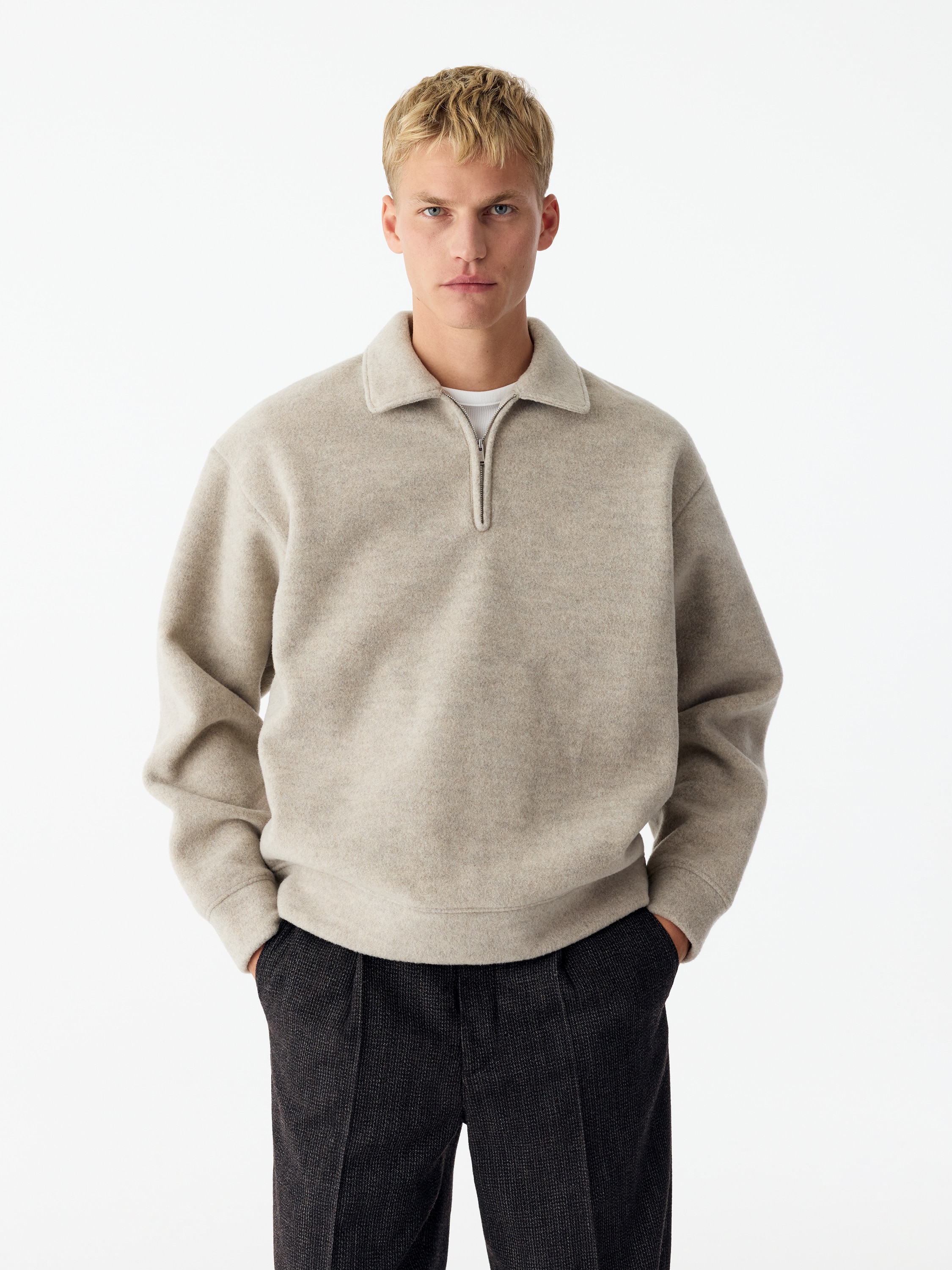 Un jersey de media cremallera en beige claro aparece de frente, con un tejido suave y texturizado, cuello abierto y cremallera metálica en tono plateado. La prenda presenta un corte relajado con hombros caÃdos, puños de canalé y bajo de canalé a juego.