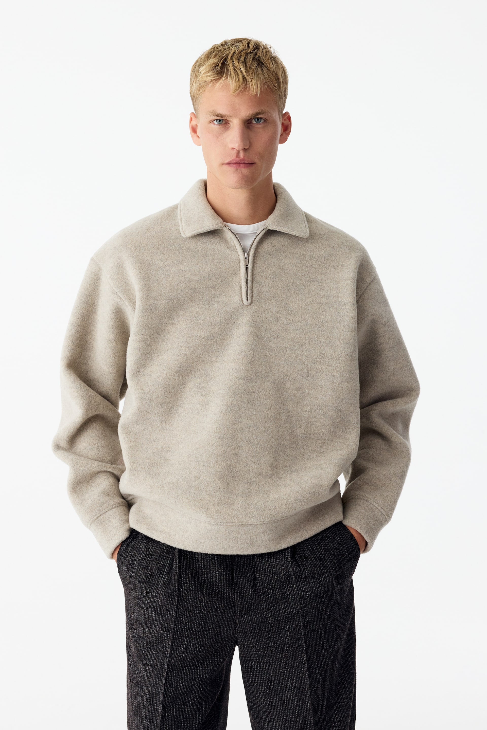 Ver imagen más grande: La modelo lleva una sudadera de media cremallera en beige claro, con textura, con cuello clásico y acabados acanalados. La sudadera se combina sobre una camiseta blanca y se estiliza con unos pantalones oscuros con textura.