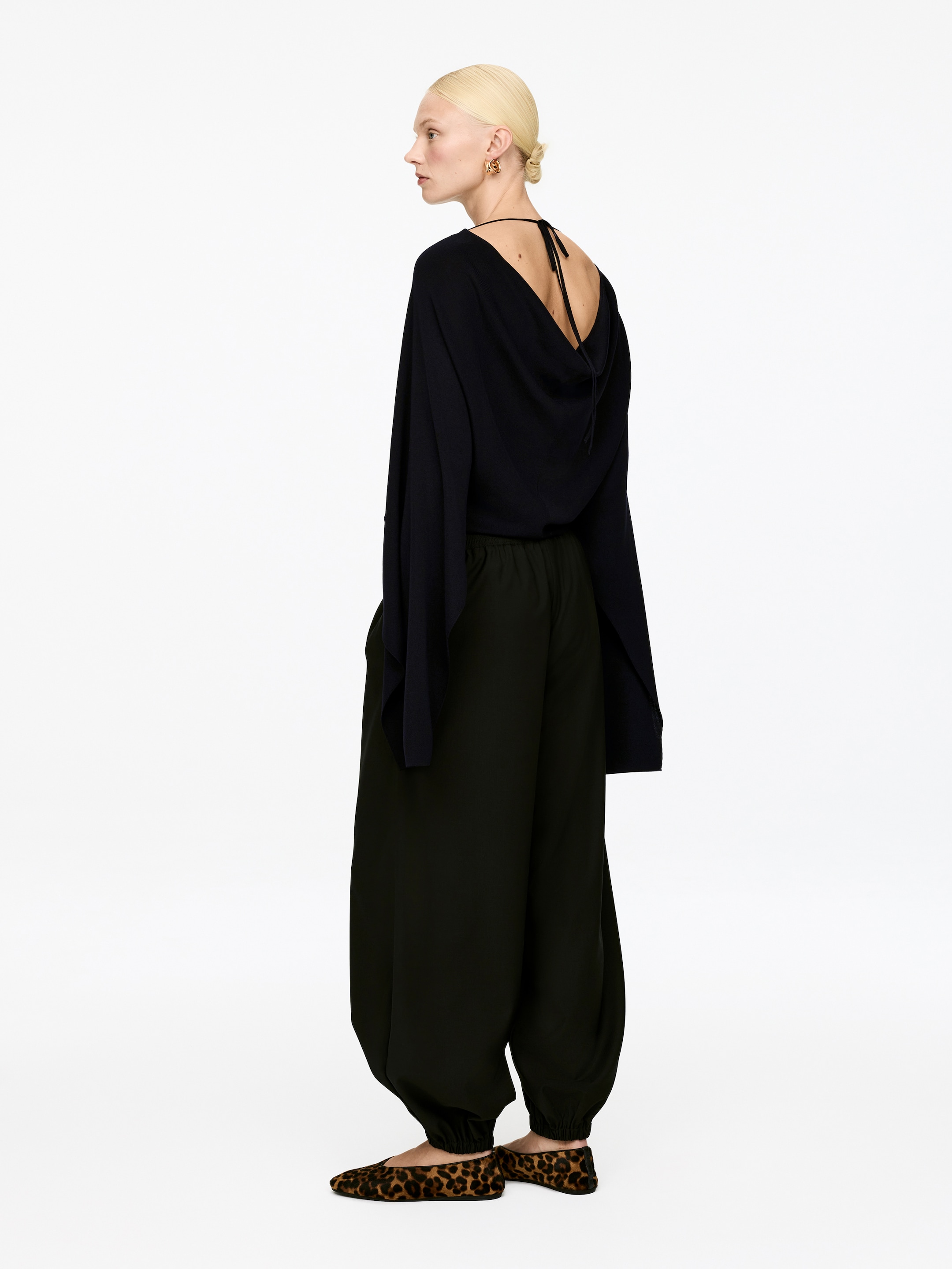Agrandir l'image: Un haut noir, présenté de dos, est doté de manches longues et larges et d'un dos drapé avec un décolleté en V et de fines attaches. Un pantalon sombre et ample, visible de côté, présente des chevilles froncées, assorti de ballerines à imprimé léopard.