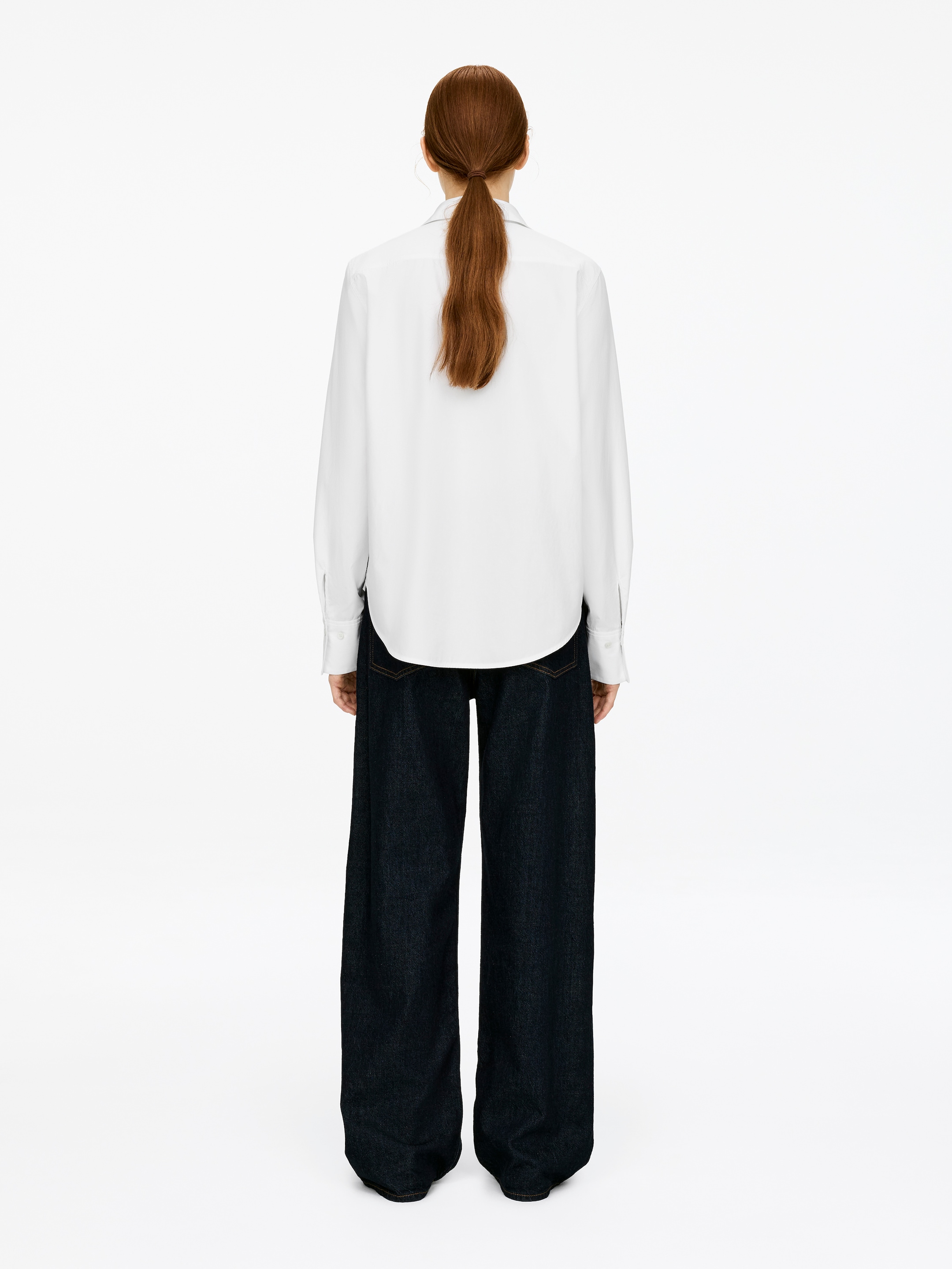 Visualizza immagine più grande: Una classica camicia di cotone è indossata dalla modella. La camicia bianca a vestibilità regolare presenta maniche lunghe con polsini e un orlo arrotondato, abbinata a pantaloni larghi scuri.