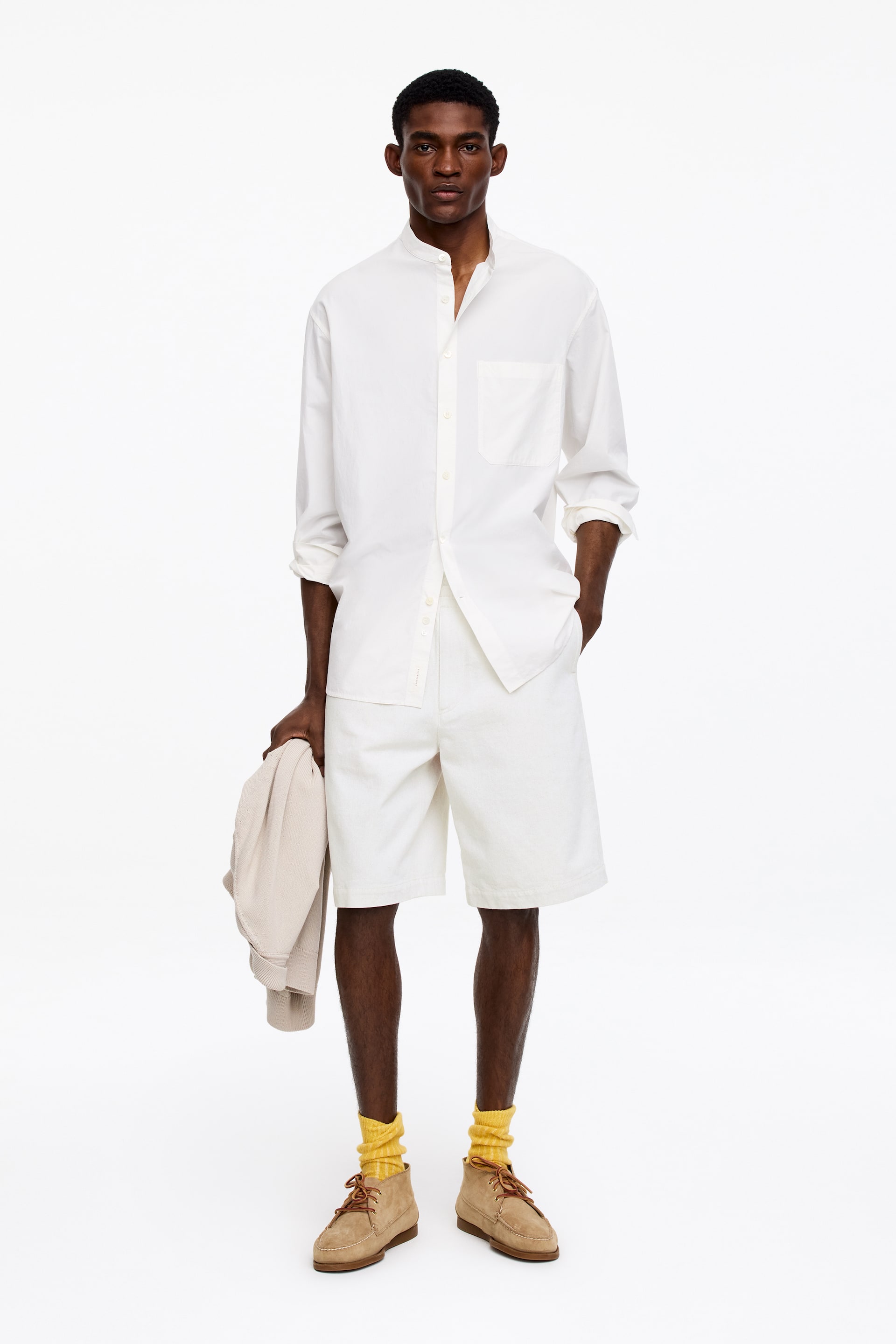 Zobraziť väčší obrázok: Biela košeľa s páskovým golierom, s ľavým náprsným vreckom a vyhrnutými dlhými rukávmi, je nosená s bielymi šortkami po kolená. Svetlobéžová bunda je držiaca v ruke a tan čukka čižmy so šnurovaním sú nosené so žltými rebrovanými ponožkami, ktoré sú nariasené.