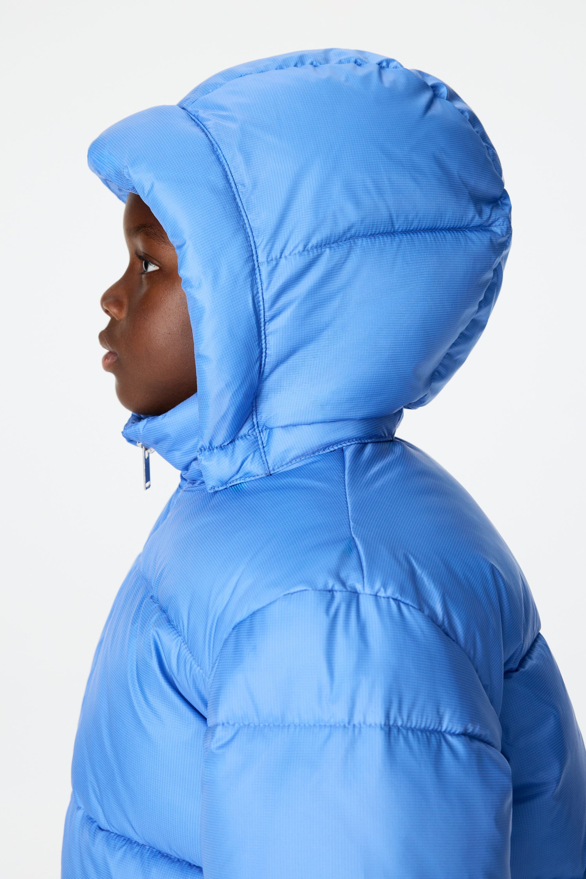 Ver imagen más grande: Una chaqueta acolchada azul se presenta de perfil lateral, con una capucha acolchada que cubre la cabeza. La chaqueta muestra una textura horizontal sutil en su tejido, y un tirador de cremallera de color plateado es visible en el cuello.