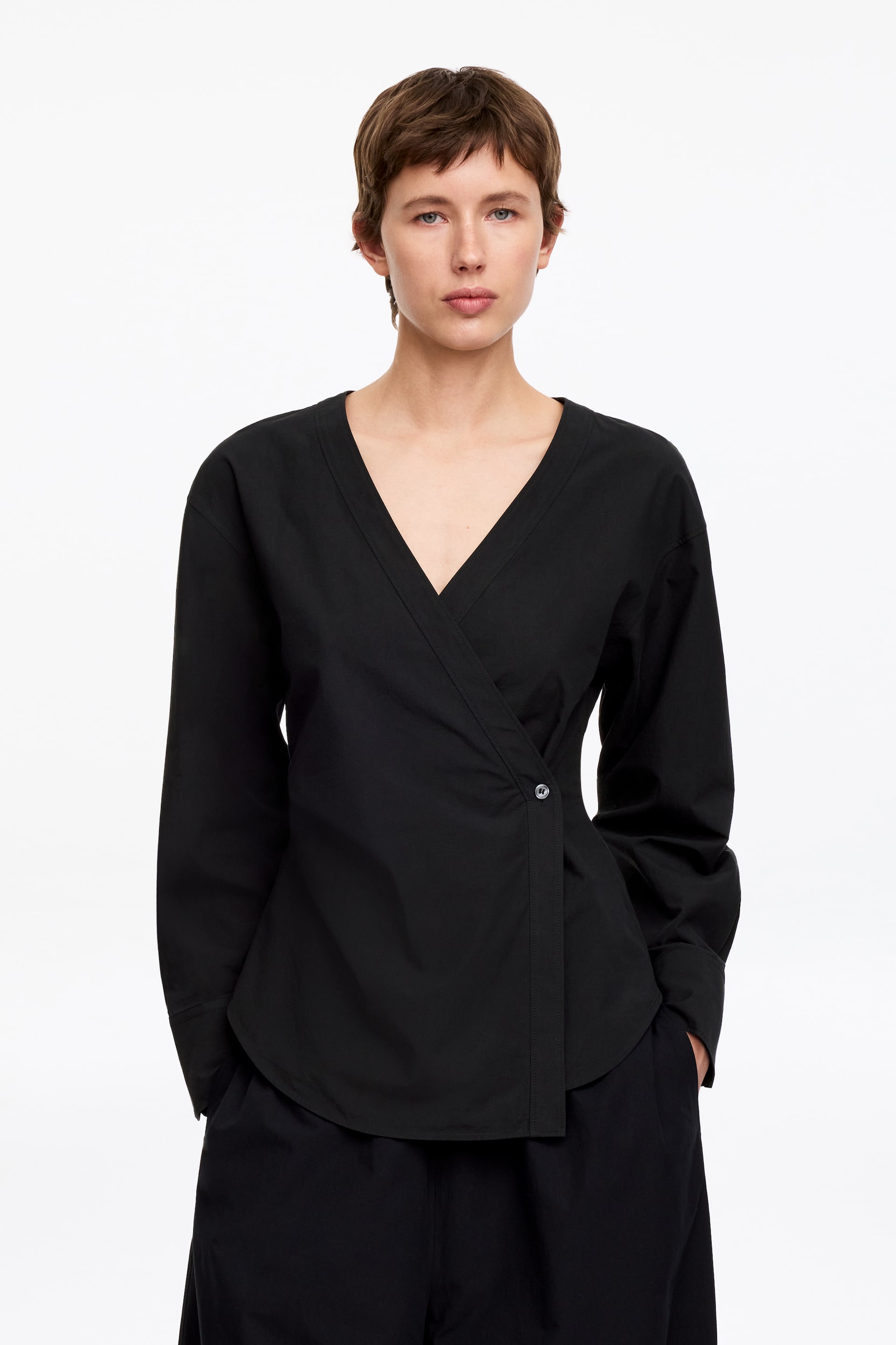 Größeres Bild anzeigen: Ein schwarzes Wickelshirt mit V-Ausschnitt und langen Ärmeln wird vom Model getragen. Es verfügt über einen seitlichen Knopfverschluss und einen abgerundeten Saum.