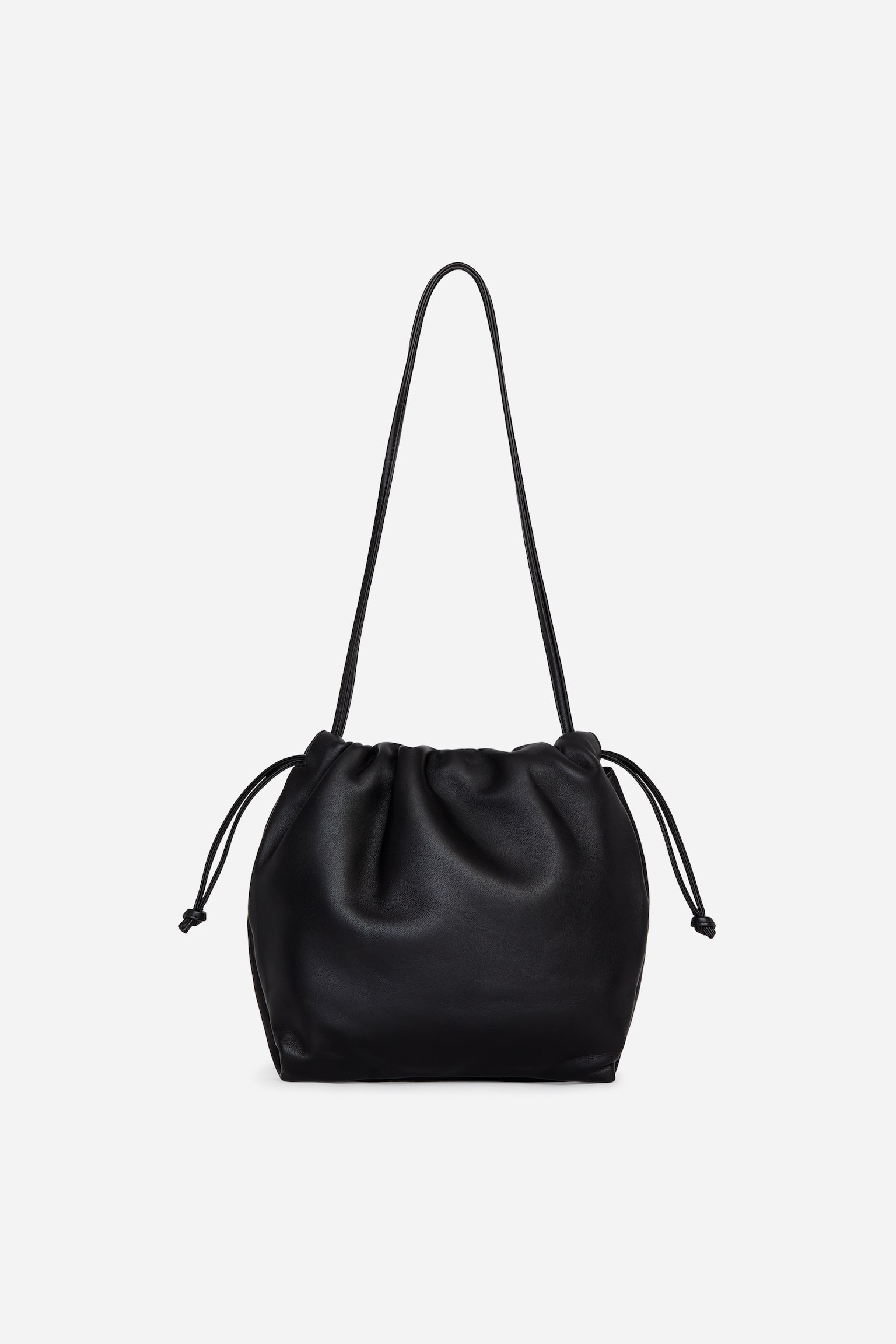 A black leather drawstring handbag is shown from the front, featuring a soft, gathered body with a ruched top closure and thin drawstrings knotted at the ends. A long, slender shoulder strap extends vertically from the top opening of the bag.