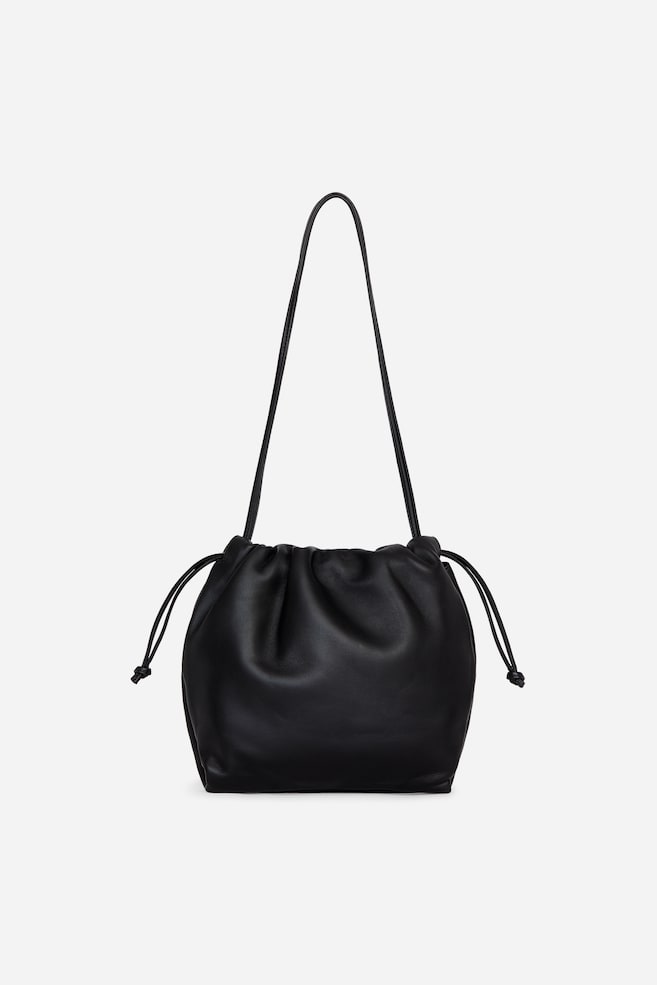 Un sac à main à cordon en cuir noir est présenté de face, mettant en valeur un corps souple et froncé avec une fermeture à cordon froncée sur le dessus et de fins liens noués à leurs extrémités. Une longue bandoulière fine sâétend verticalement depuis lâouverture supérieure du sac. - 1