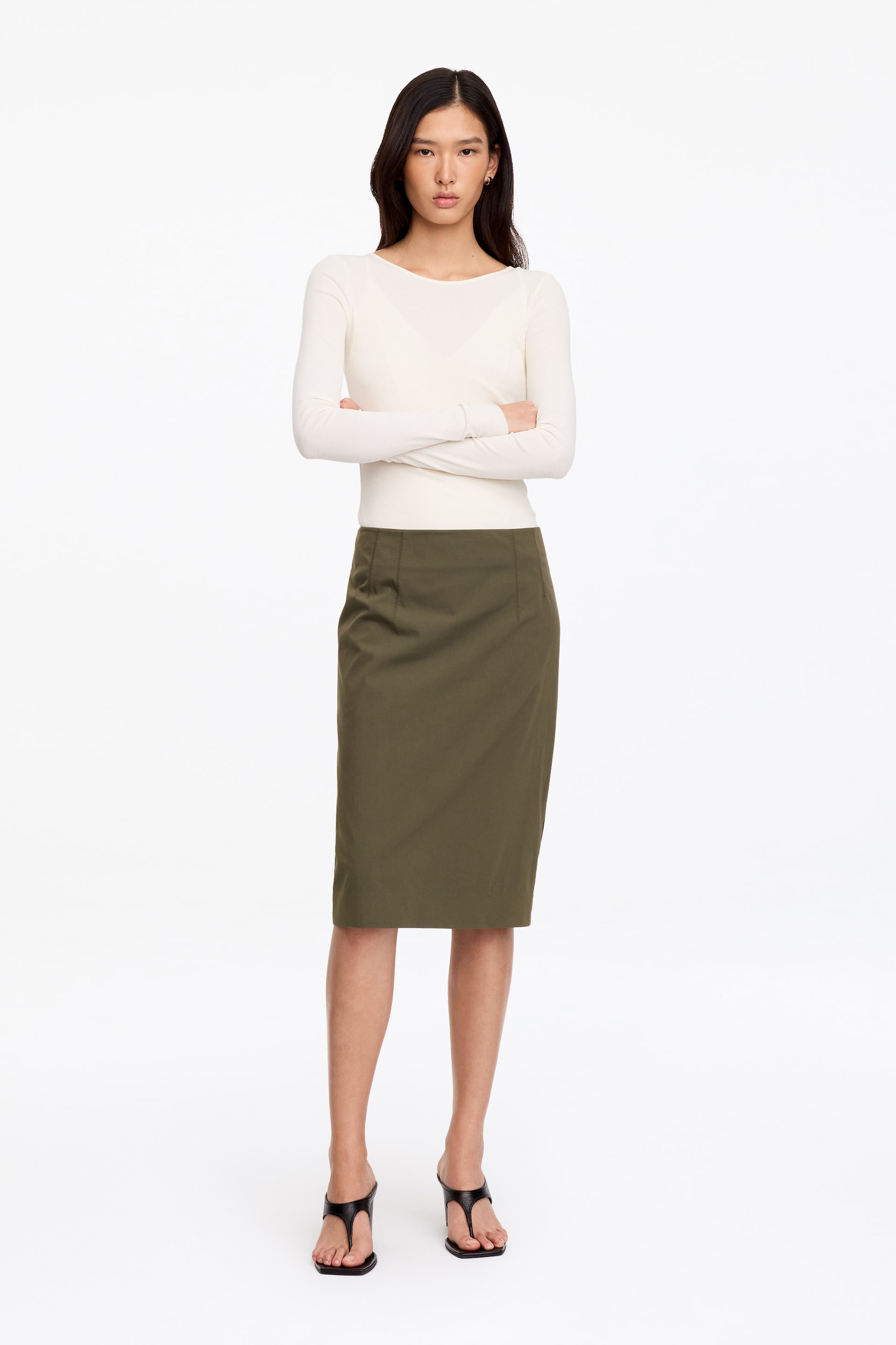 Visualizza immagine più grande: La modella indossa una gonna a matita verde oliva, con vita media e una silhouette strutturata al ginocchio. La gonna è abbinata a un top color bianco sporco a maniche lunghe infilato all’interno.
