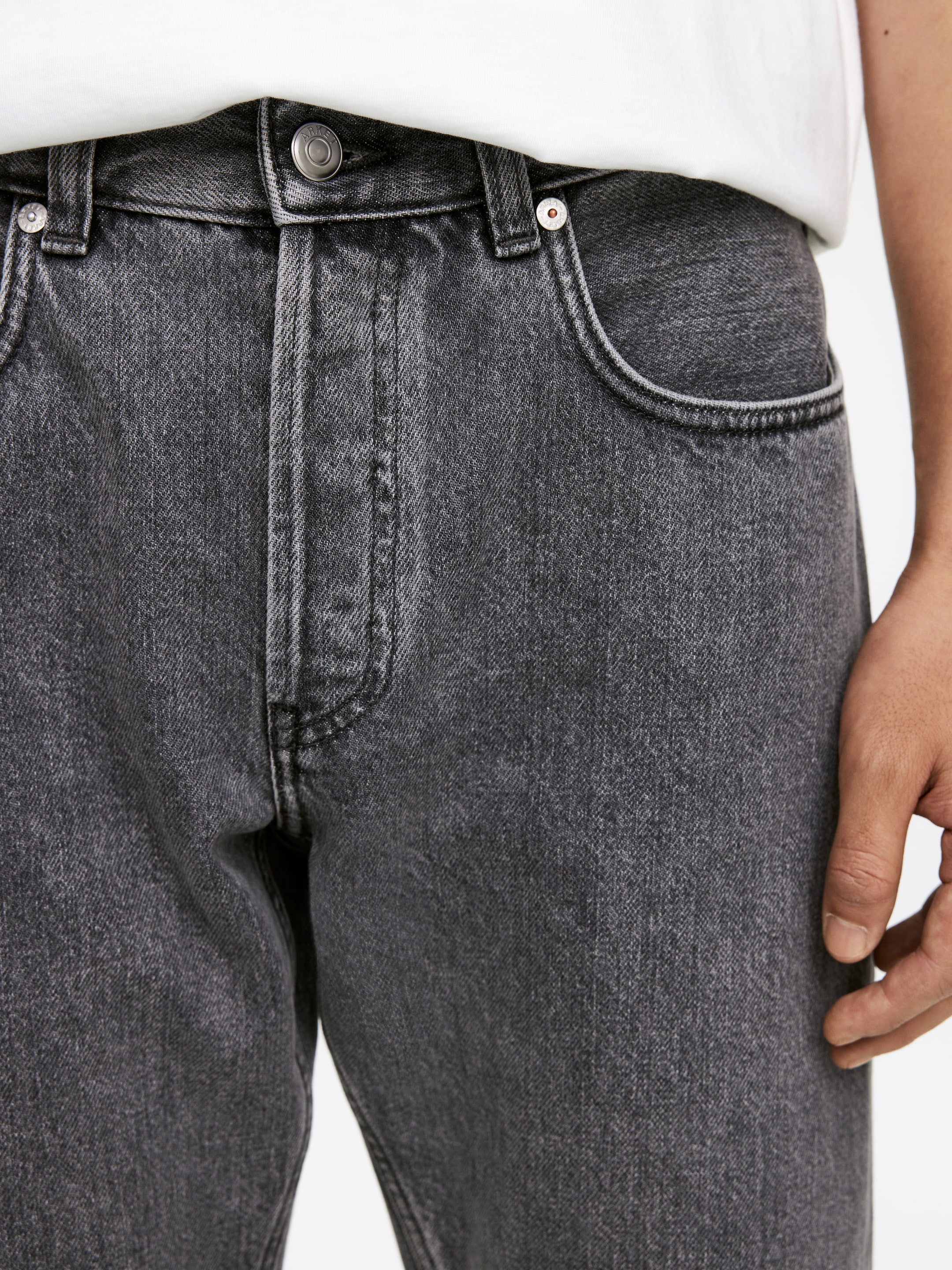 Grösseres Bild ansehen: Dunkelgraue Denim-Jeans werden von vorne präsentiert, mit einer verwaschenen Optik, einem sichtbaren Knopfverschluss mit einem Metallknopf und einem Bund, der Gürtelschlaufen zeigt. Abgerundete Vordertaschen werden ebenfalls gezeigt, von denen eine ein Metallniete hervorhebt, während ein weißes T-Shirt in den Bund gesteckt getragen wird.