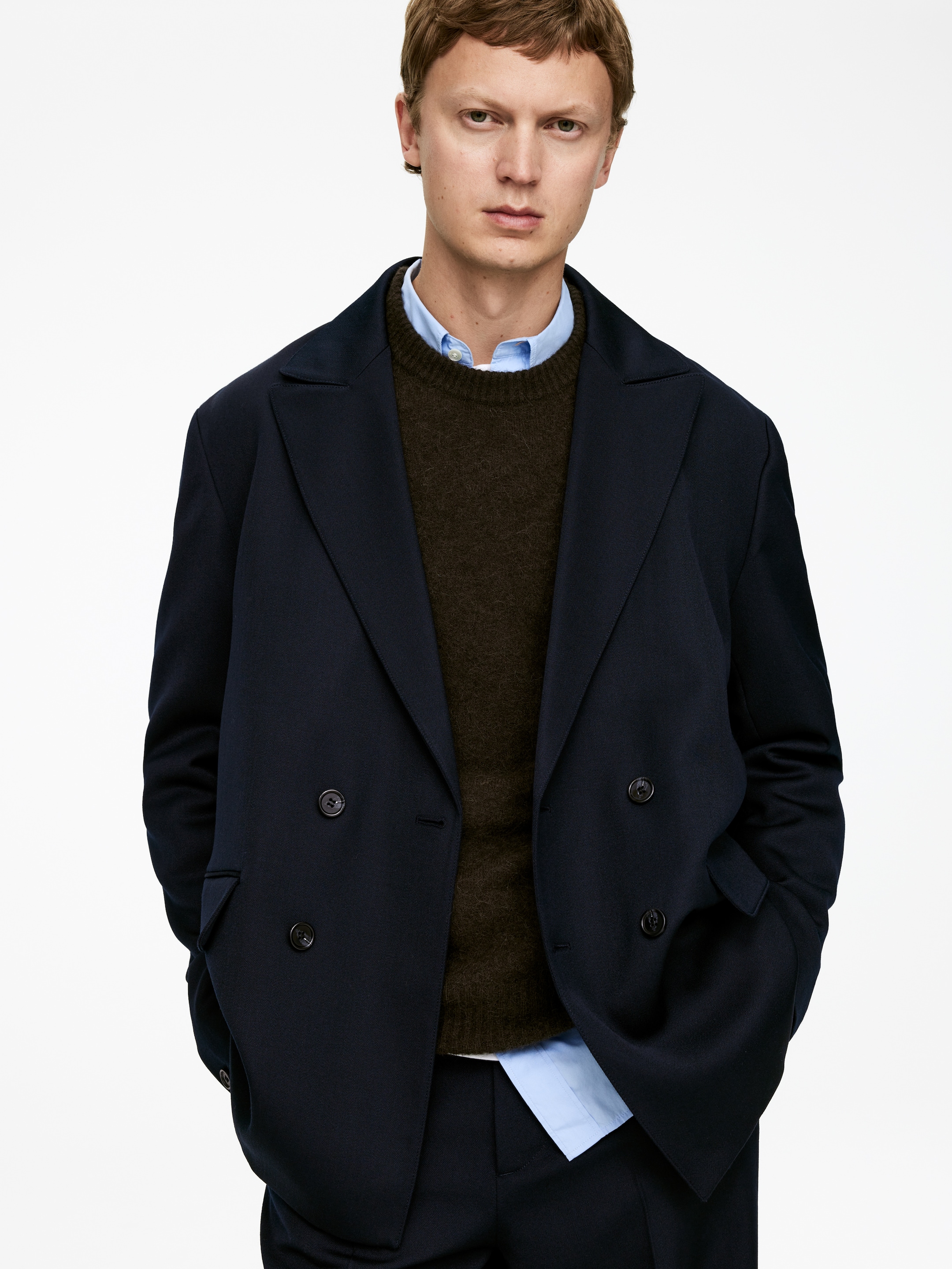 Größeres Bild ansehen: Ein marineblauer zweireihiger Blazer wird aus einem frontalen Winkel getragen, mit breiten Kerbrevers, zwei Reihen dunkler Knöpfe und zwei aufgesetzten Taschen. Er ist über einem dunkelbraunen Rundhalspullover und einem hellblauen Hemd mit Kragen geschichtet, wobei passende marineblaue Hosen ebenfalls sichtbar sind.