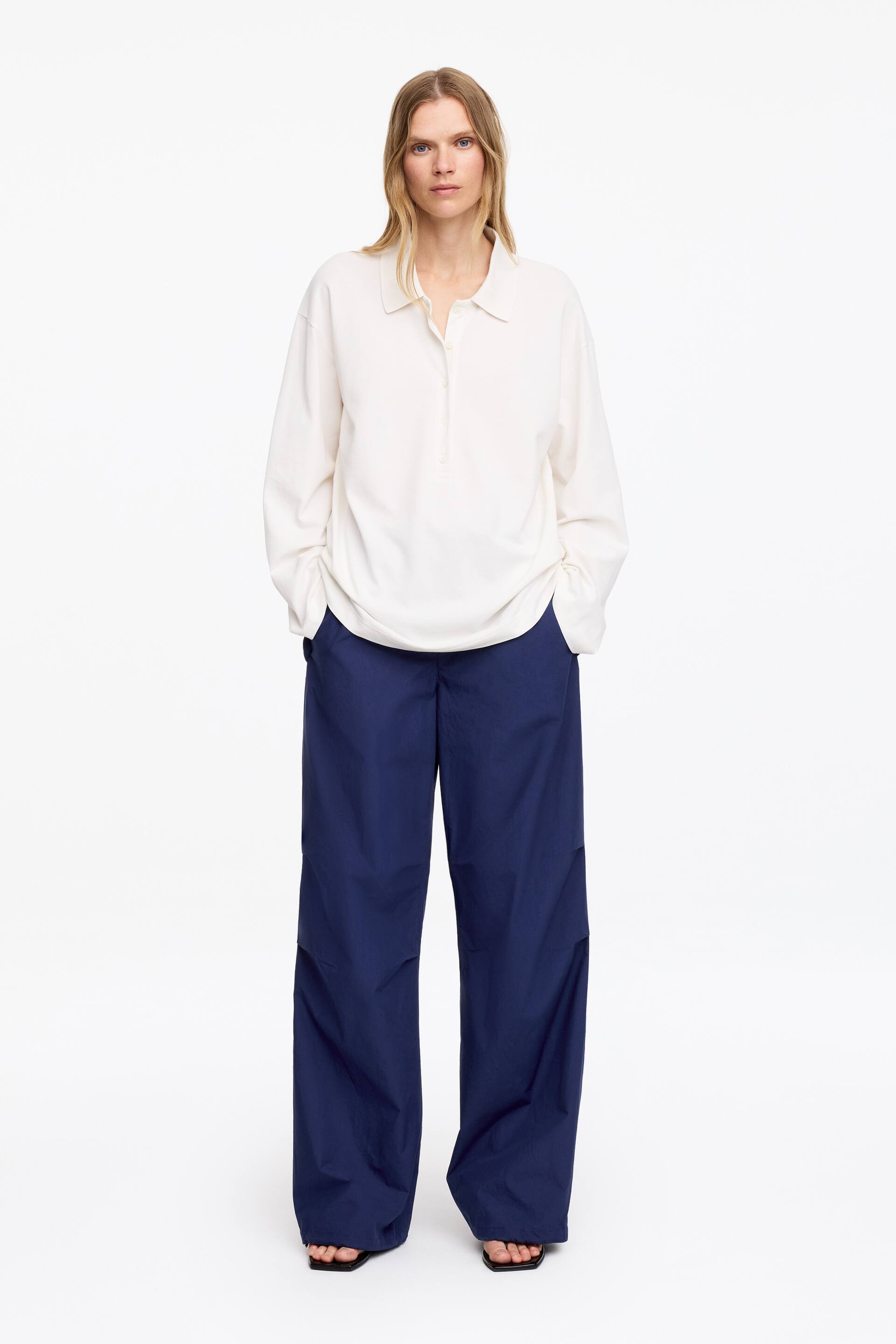 Visualizza l'immagine ingrandita: Una polo a maniche lunghe color avorio, con colletto e abbottonatura parziale, è indossata con pantaloni blu scuro a gamba larga che cadono con un drappeggio rilassato, rivolti in avanti.