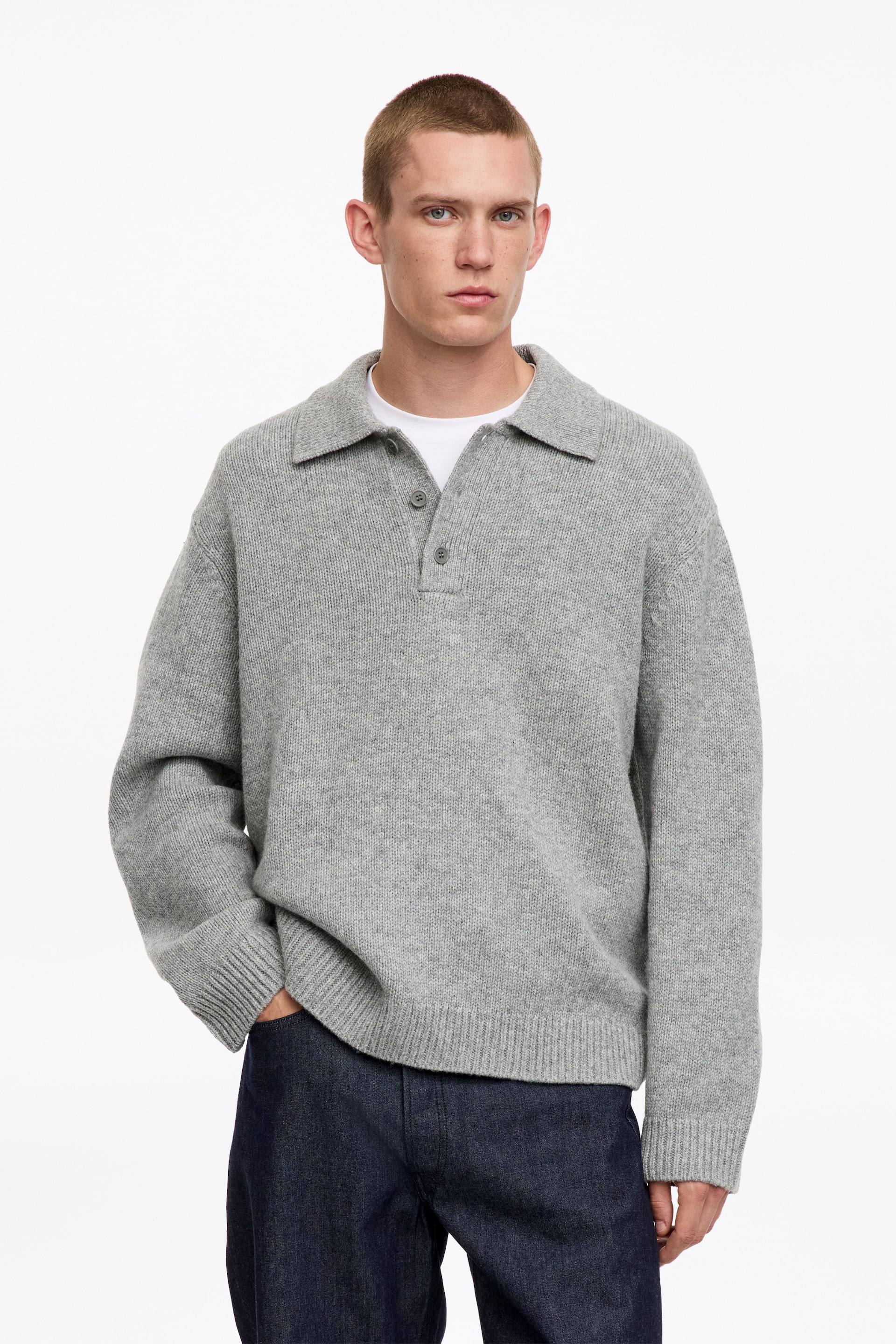 Visualizza immagine più grande: La modella indossa un maglione polo in lana lavorata a maglia grigio chiaro. Presenta colletto polo, mezza abbottonatura a tre bottoni e vestibilità rilassata, indossato sopra una t-shirt bianca. La modella è in piedi con una mano appoggiata nella tasca dei pantaloni.
