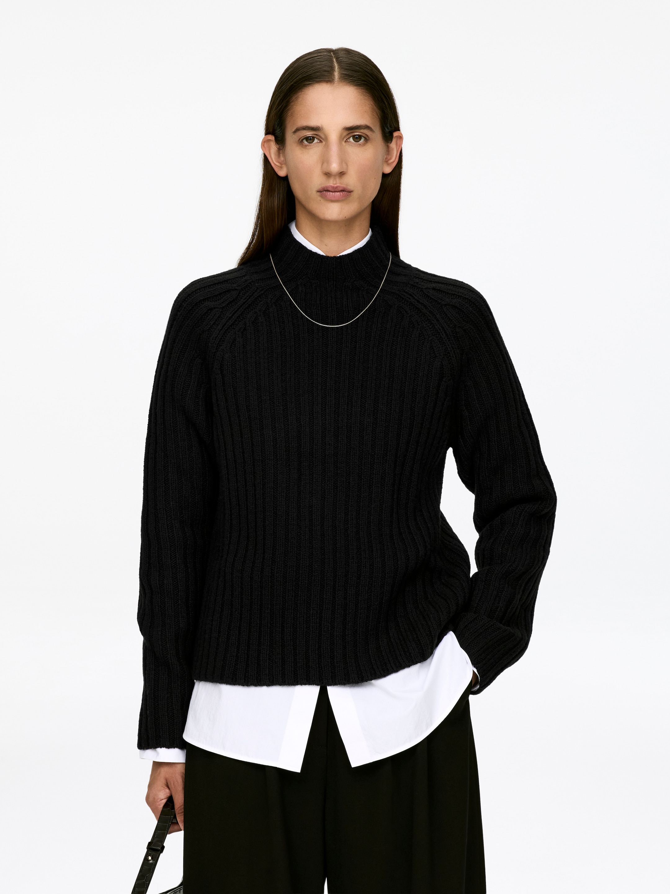 Agrandir l'image: Un pull noir en maille côtelée avec un col montant et des manches raglan longues est porté par le mannequin, superposé à une chemise blanche à col dont le col et l'ourlet sont visibles. Un collier fin en argent repose sur le col du pull, et le mannequin tient un sac sombre.