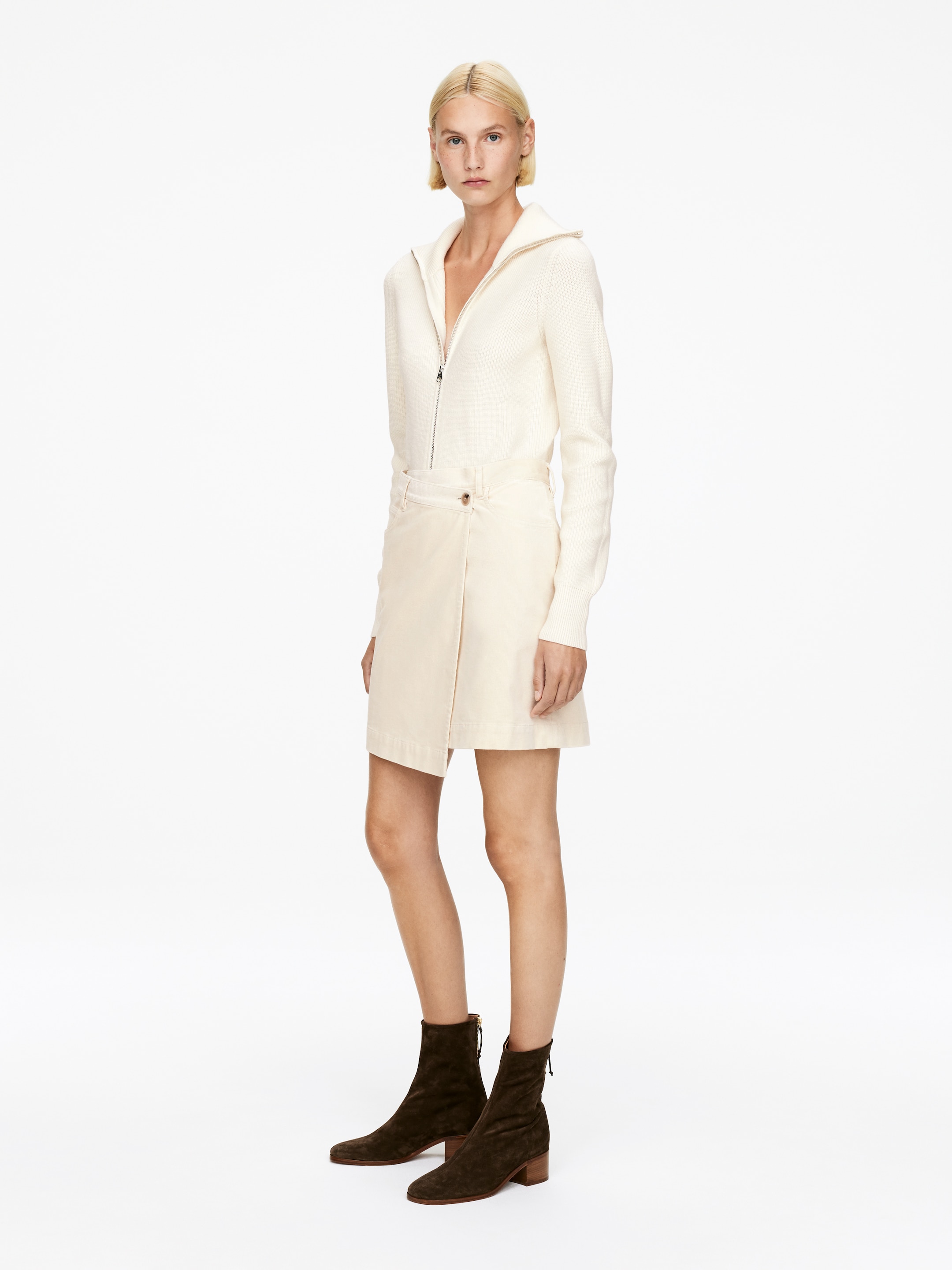 Vis større bilde: Et off-white antrekk blir presentert fra en liten vinkel, og viser frem en ribbestrikket full-zip topp med ståkrage som er åpnet til en V-hals og ribbestrikkede mansjetter, sammen med et omslagsskjørt som er festet med en knapp på venstre hofte. Mørkebrune ankelstøvletter i semsket skinn med blokkhæler og glidelåser bak fullfører antrekket.