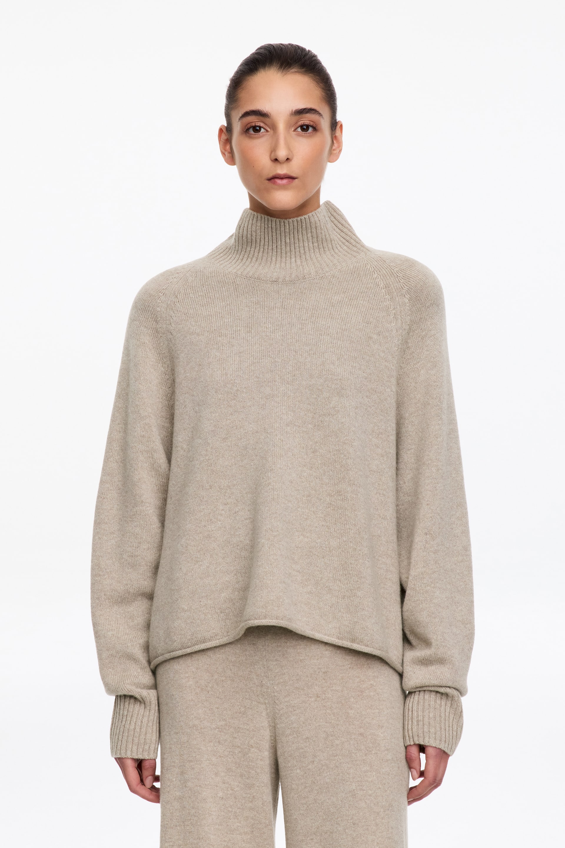 Größeres Bild anzeigen: Ein beigefarbener Strickpullover wird vom Model getragen und zeichnet sich durch einen hoch geschnittenen Rippkragen, Raglanärmel mit langen Rippbündchen und eine lockere Passform aus. Der Pullover ist mit einer dazu passenden Strickhose in derselben Farbe und Textur kombiniert.