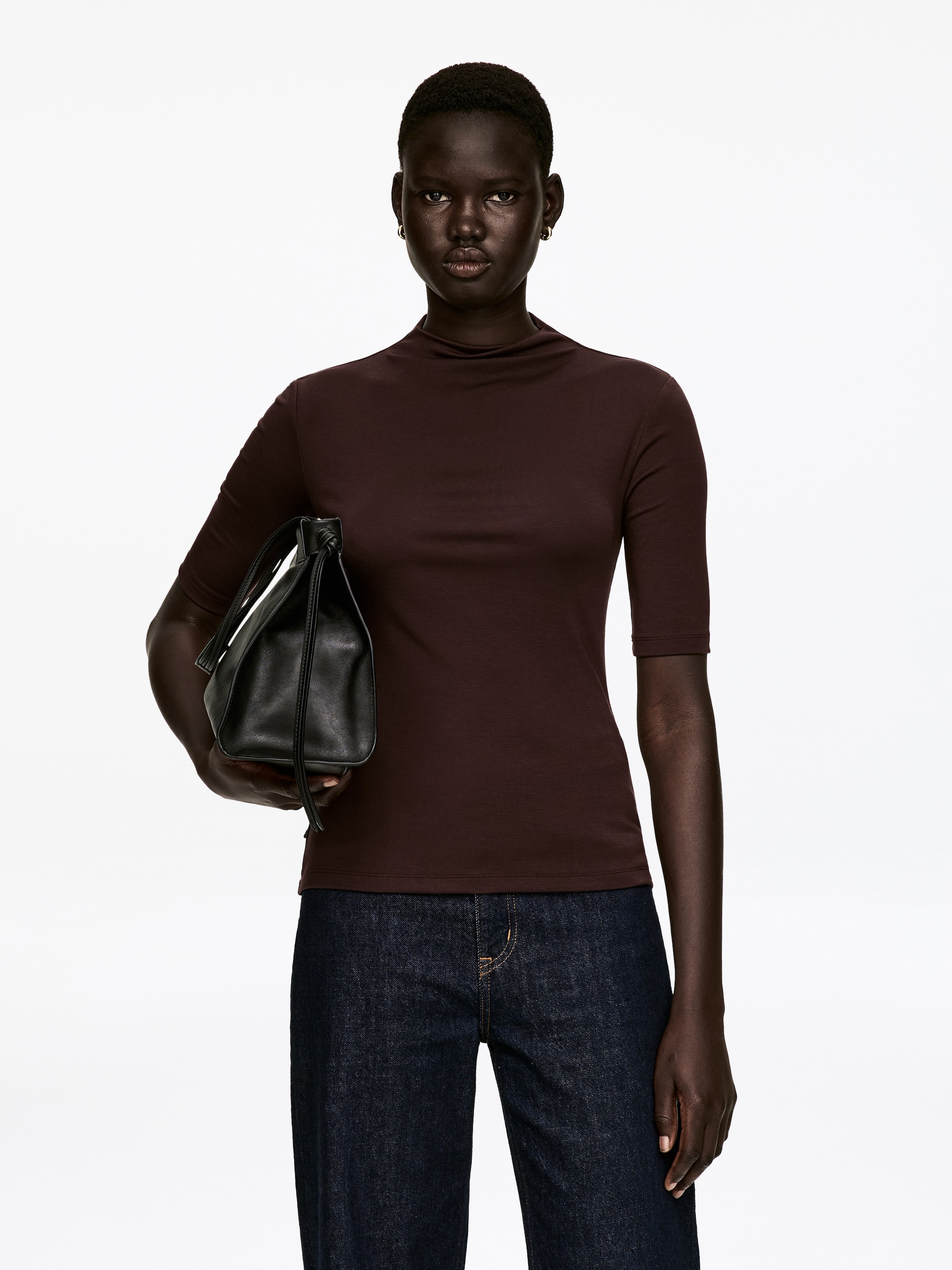 Преглед на по-голямо изображение: Тъмнокафяво поло с висока яка е представено отпред, с плътно прилепване и ръкави до лактите. То демонстрира гладка текстура и завършва на талията, комбинирано с тъмни дънки.