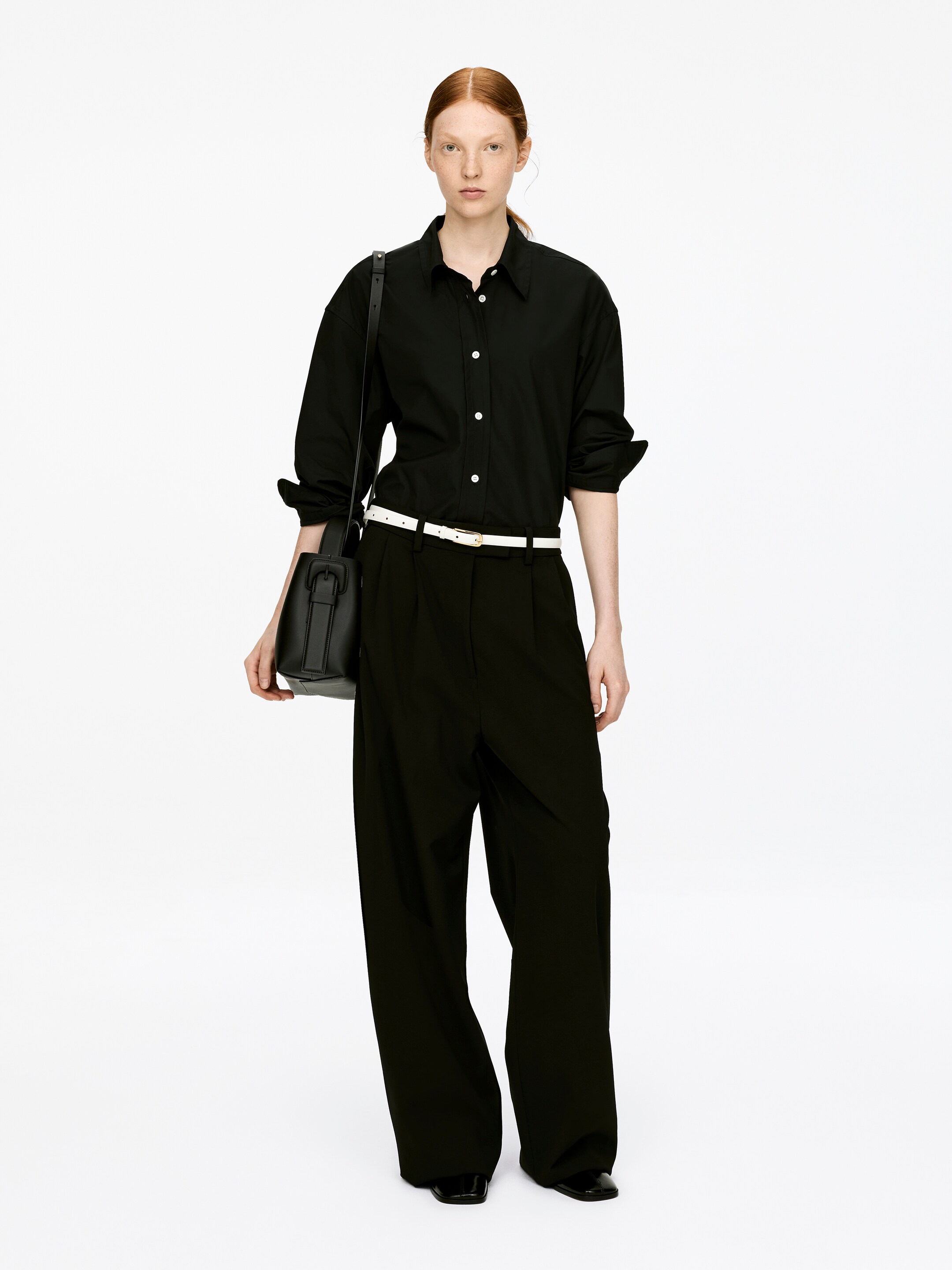 View larger image: A structured black mid-size crossbody bag is being carried by the model on their left shoulder, its adjustable strap crossing the body. The bag's wide top handle, with a buckle detail, is being held by the model's left hand.
