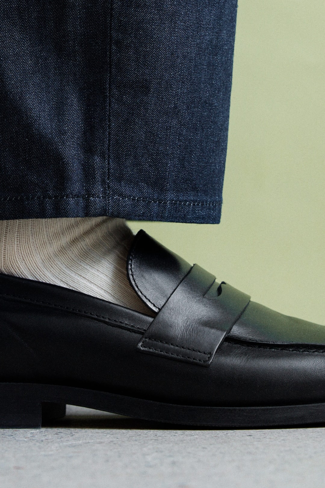 Ver imagen más grande: Un mocasín de cuero negro tipo "penny loafer" se lleva puesto, con una tira sobre el empeine y una punta redondeada, presentado en un perfil lateral. Se combina con un calcetín ligero de canalé y unos vaqueros oscuros que descansan sobre el zapato.