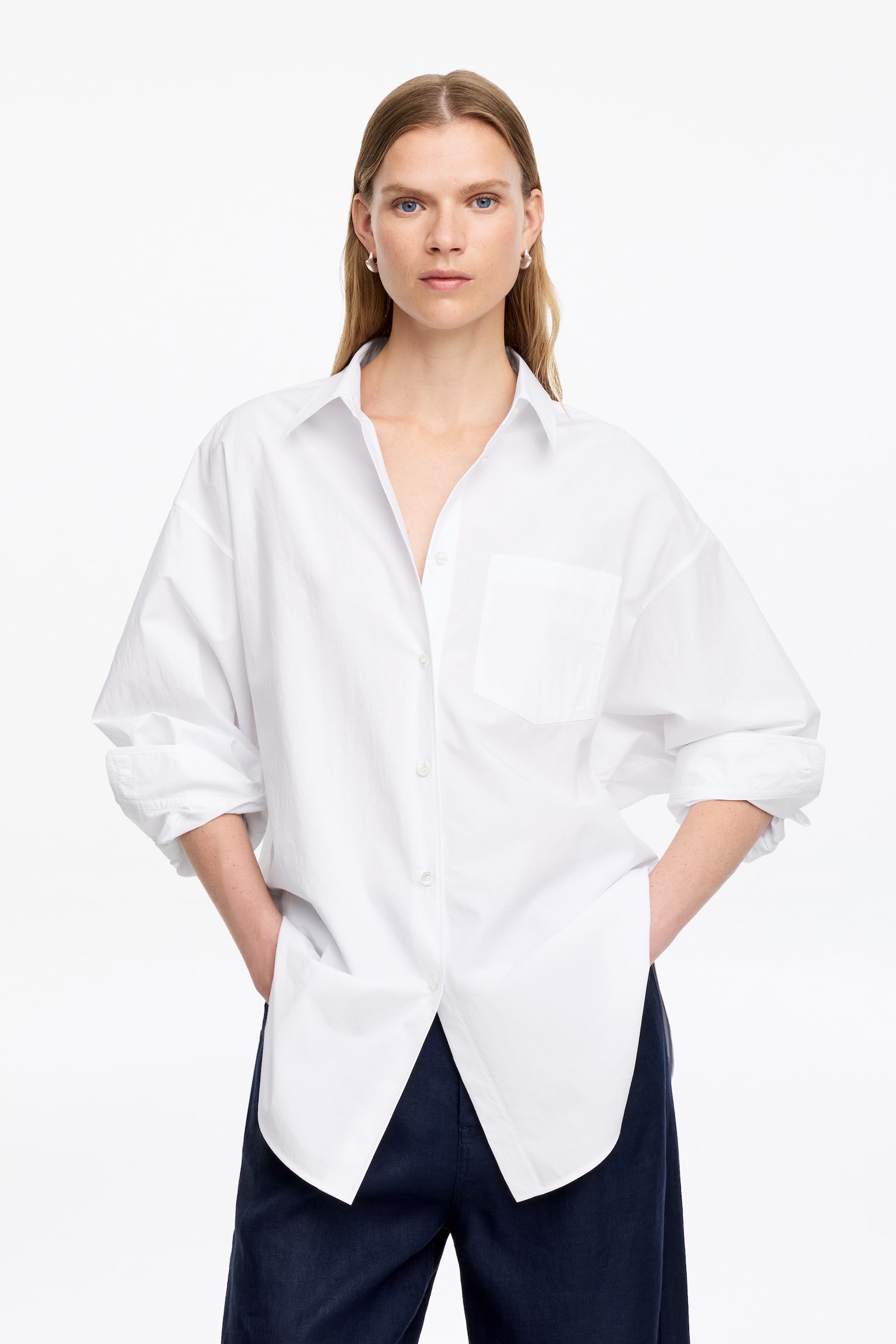 Ein weißes Button-Down-Hemd mit offenem, spitzem Kragen und teilweise geöffnetem Knopfverschluss. Es präsentiert eine aufgesetzte Brusttasche auf der linken Seite, lange Ärmel, die bis zu den Unterarmen hochgekrempelt sind, eine lockere Passform und einen abgerundeten Saum, der lässig über dunklen Hosen hängt. - 1