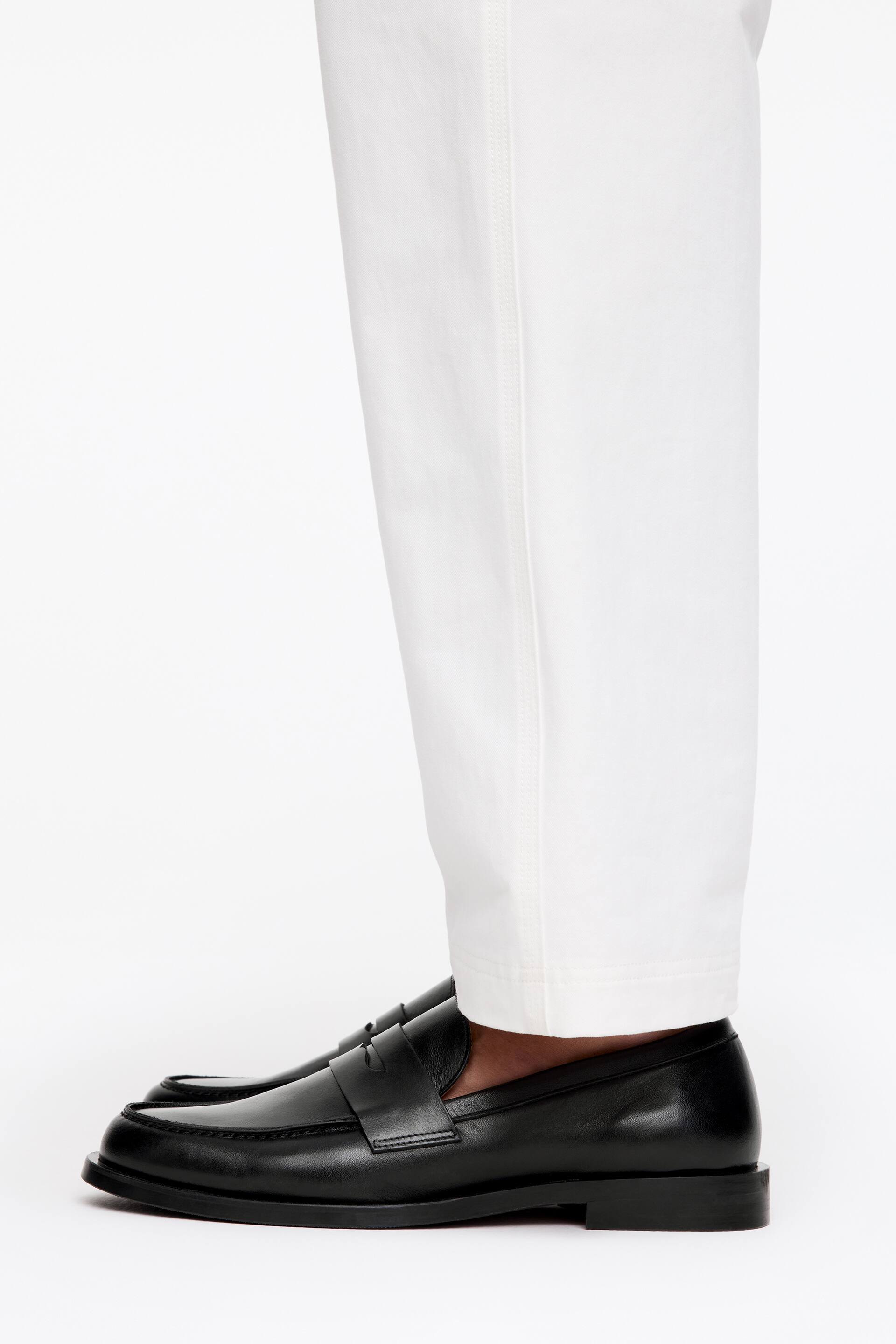 Grotere afbeelding bekijken: Zwarte semi-glanzende leren loafers, met een gestapelde zool en welt-details, worden gedragen door het model, wiens witte broek net boven de schoenen rust.