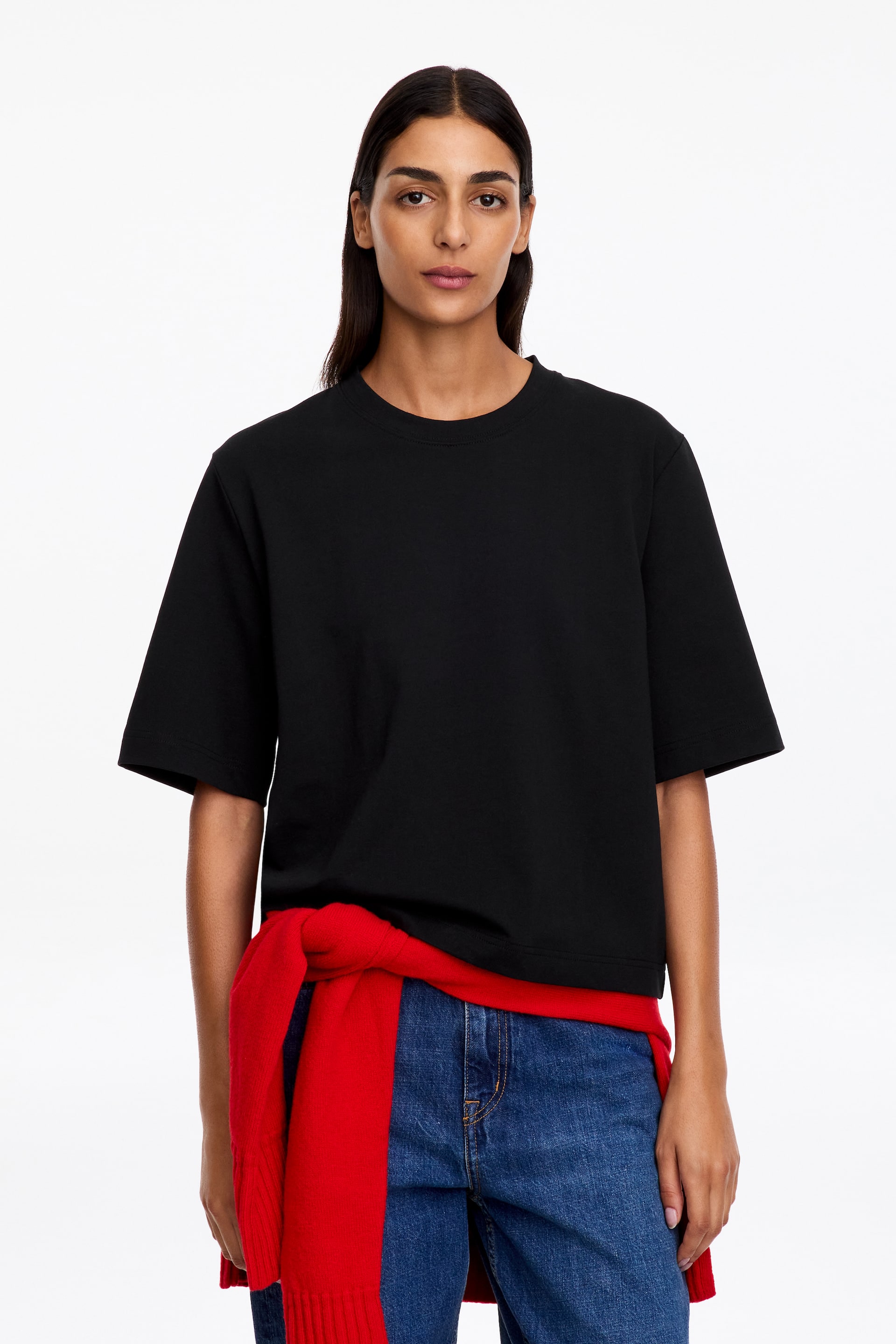Agrandir l'image: Un T-shirt noir en coton, à la coupe boxy, aux épaules tombantes et aux manches courtes allongées, est porté par le mannequin. Ce style légèrement court est associé à un jean bleu et à un pull rouge noué autour de la taille.
