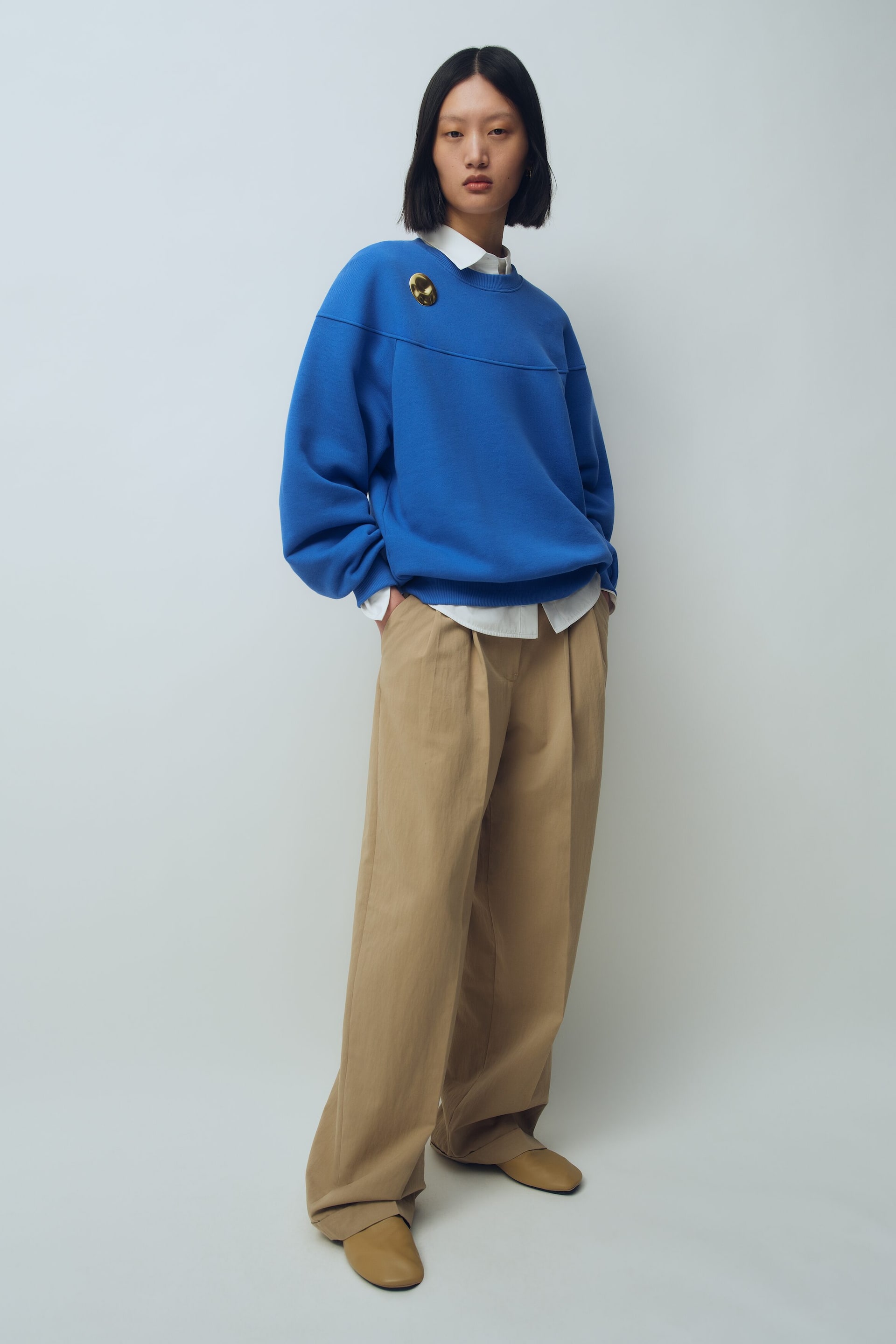 Visa större bild: En klarblå sweatshirt med rund halsringning, avslappad passform, nedsänkta axlar och en guldbrosch bärs lager på lager över en vit skjorta med krage. Outfiten kompletteras med vida, veckade beige byxor som faller löst över platta skor i tan, visade i en trekvartsvinkel.
