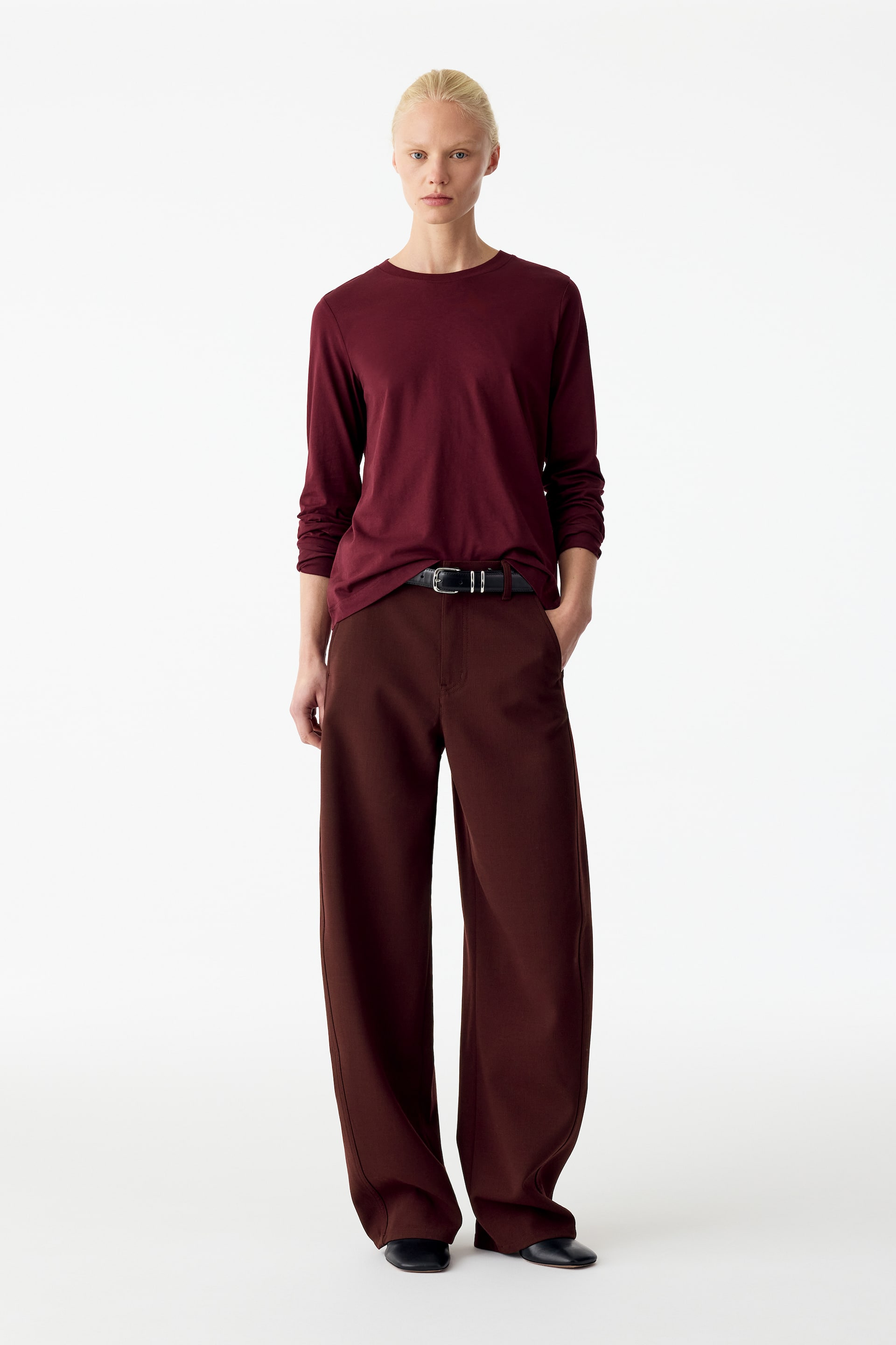 Ver imagen más grande: El modelo lleva unos pantalones marrones de mezcla de lana con una silueta relajada de pernera curvada tipo barrel, posando de pie con una mano apoyada en el bolsillo lateral. Los pantalones se combinan con un cinturón de cuero negro, un top burdeos de manga larga y unos zapatos planos negros.
