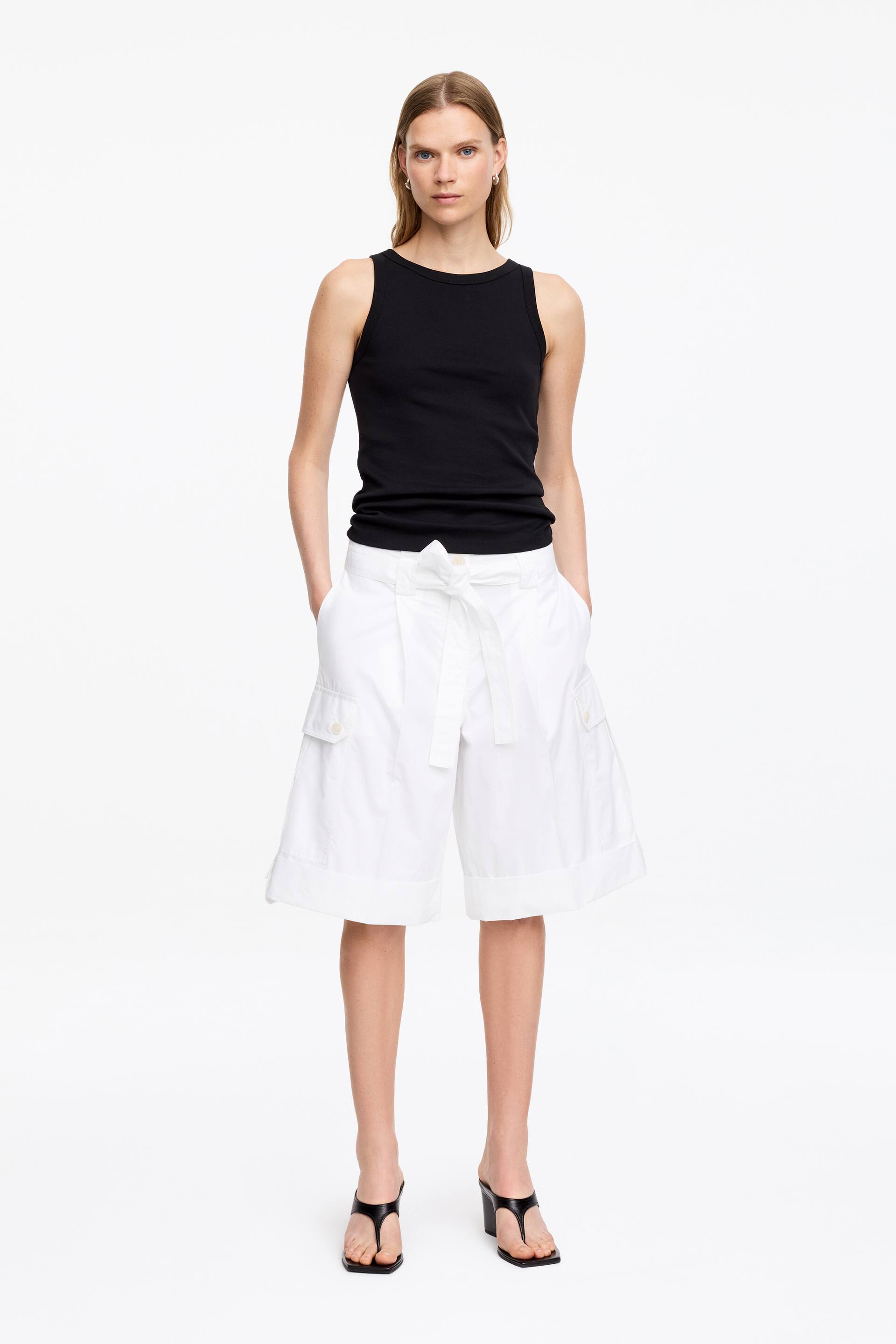 Agrandir l'image: Un débardeur noir est porté, avec un col rond et une silhouette ajustée. Il est associé à un short blanc à jambes larges, cintré à la taille par une ceinture à nouer à l'avant et doté de poches cargo boutonnées sur les cuisses, s'étendant juste au-dessus du genou.