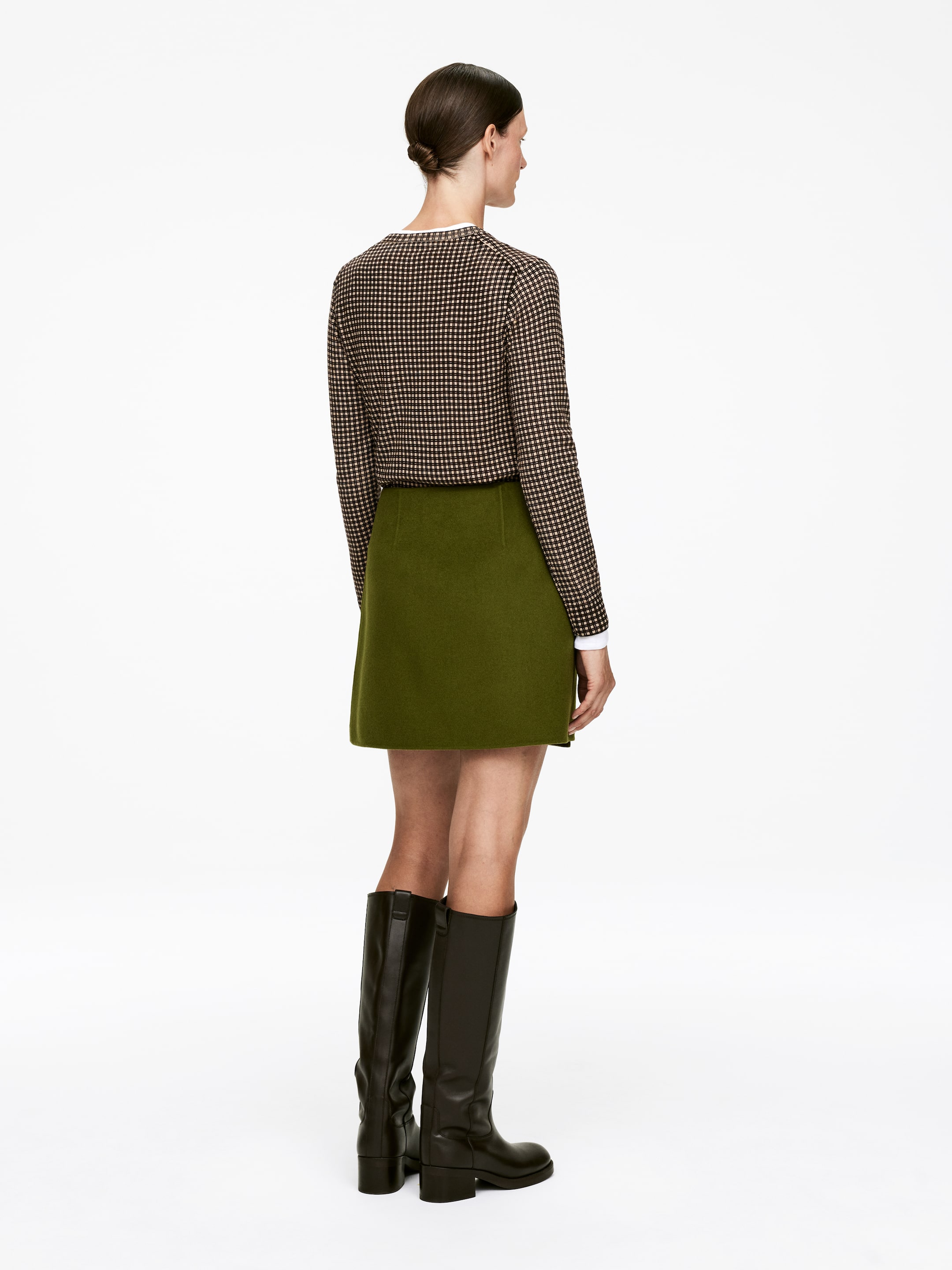 Se større billede: Et outfit med en langærmet, brun og off-white småternet top, lagdelt over en hvid skjorte med krave, vises bagfra, parret med en olivengrøn A-formet mininederdel og høje, mørkebrune læderstøvler.
