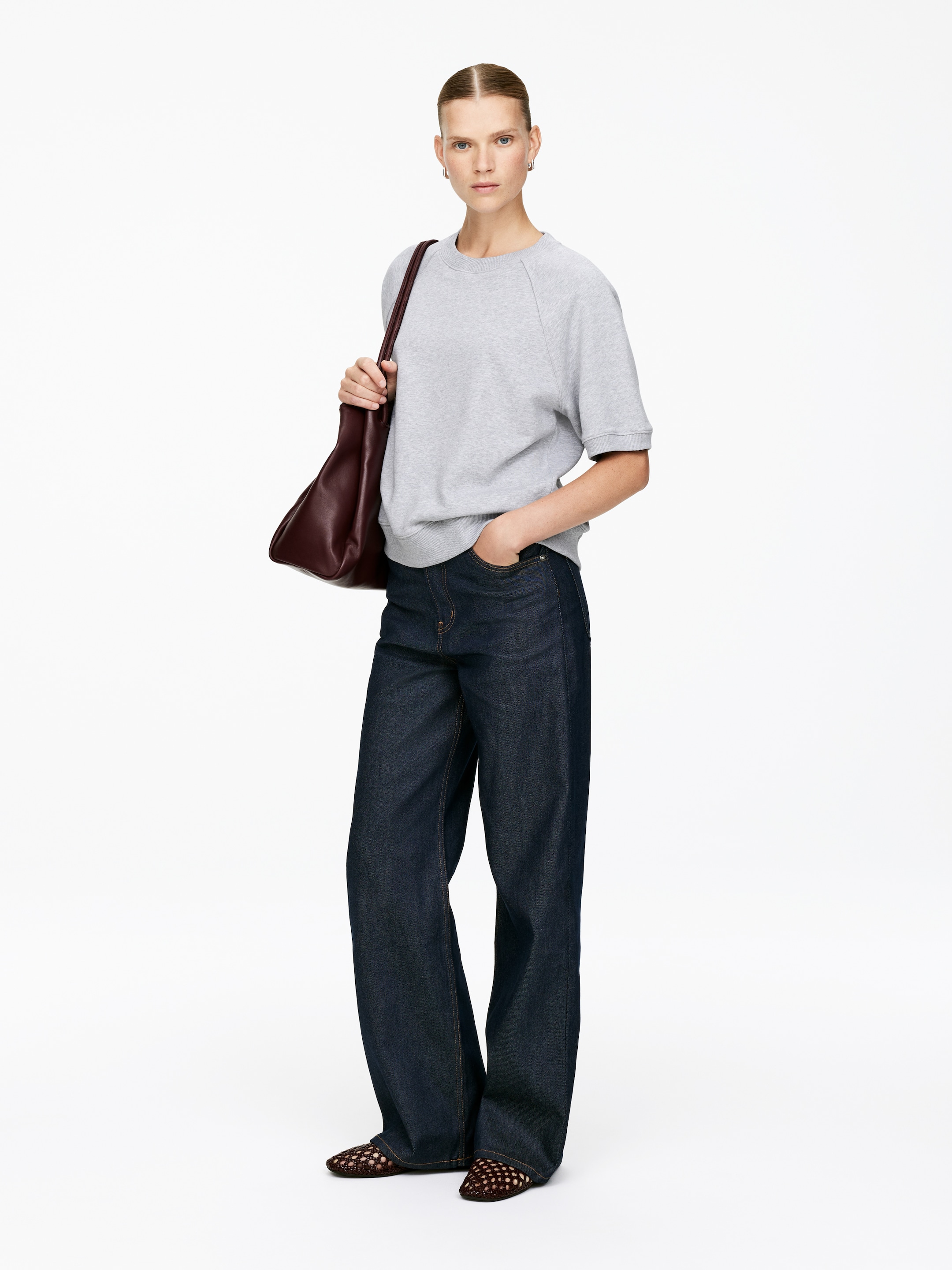 Agrandir l'image: Un sweat-shirt gris à manches courtes avec des manches raglan et un ourlet côtelé est porté avec un jean large à délavage foncé. Un sac à bandoulière bordeaux décontracté est porté, et des chaussures plates tissées foncées sont visibles.
