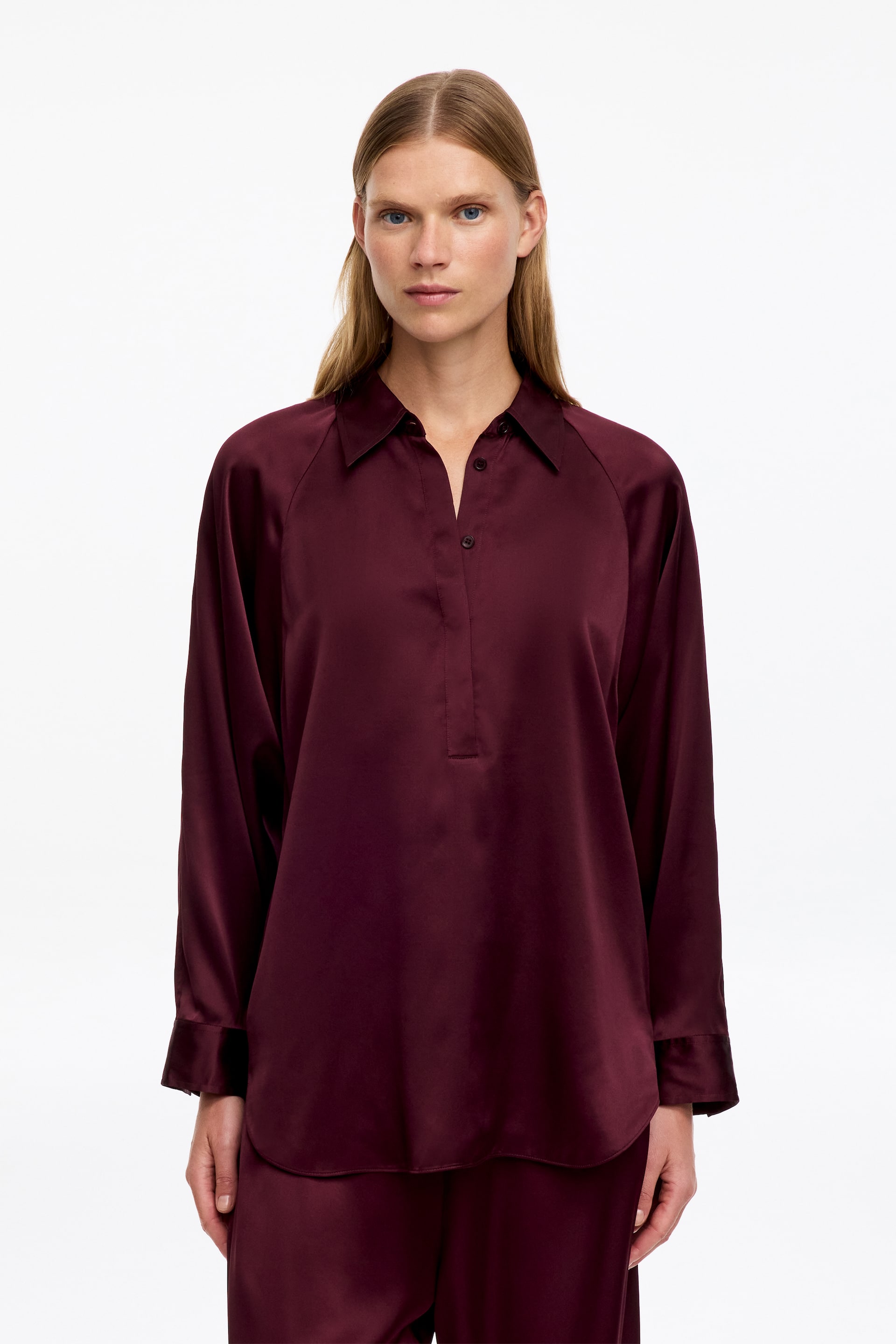 Visualizza immagine più grande: La modella indossa una camicia a maniche lunghe in raso color bordeaux intenso, con colletto a punta, abbottonatura parziale e orlo arrotondato. La camicia è abbinata a pantaloni in raso bordeaux coordinati.