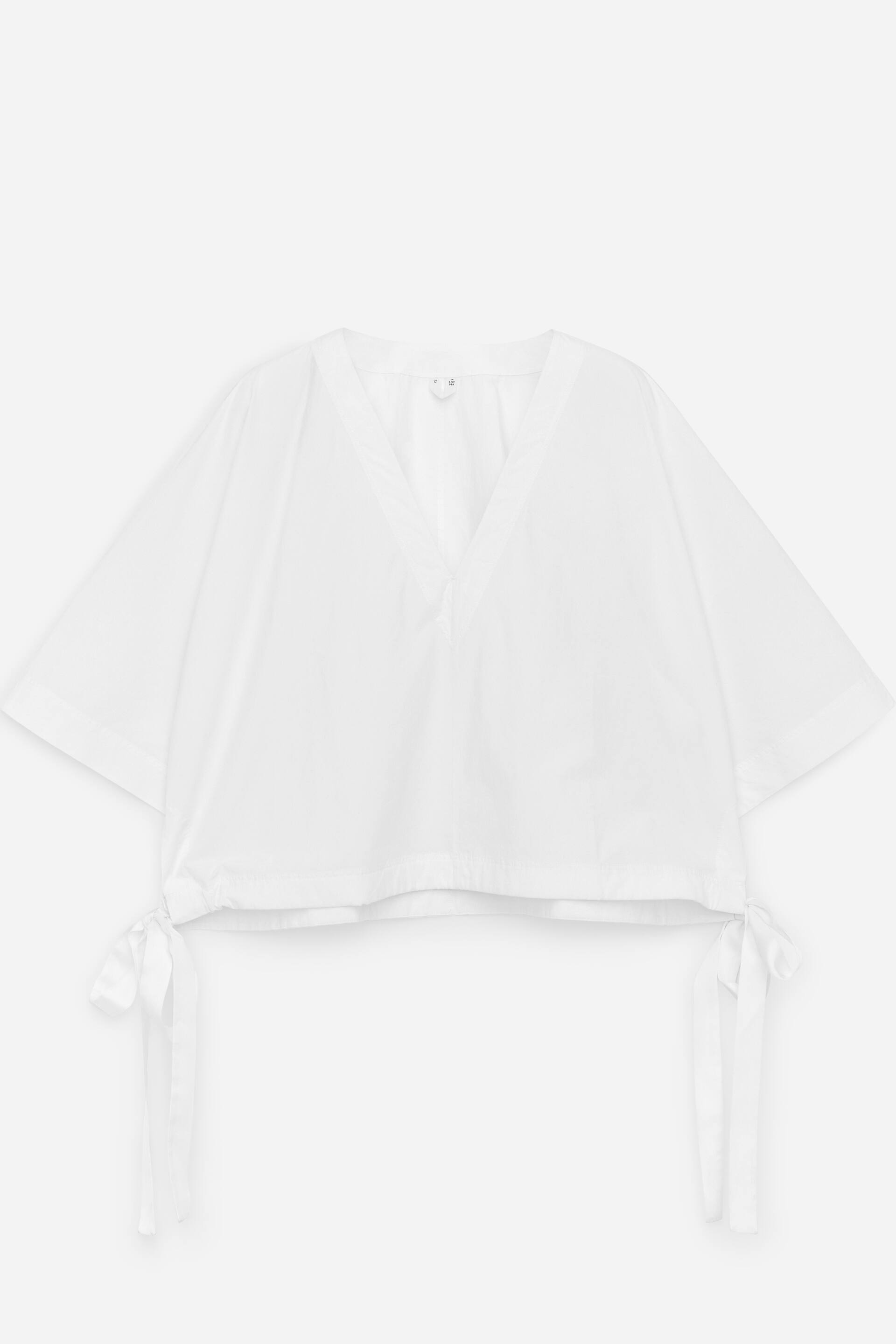 Zobacz większy obraz: Biały top przedstawiony z przodu, z dekoltem w serek i krótkimi, szerokimi rękawami. Krótki dół posiada sznurek ściągający z widocznymi wiązaniami po bokach.