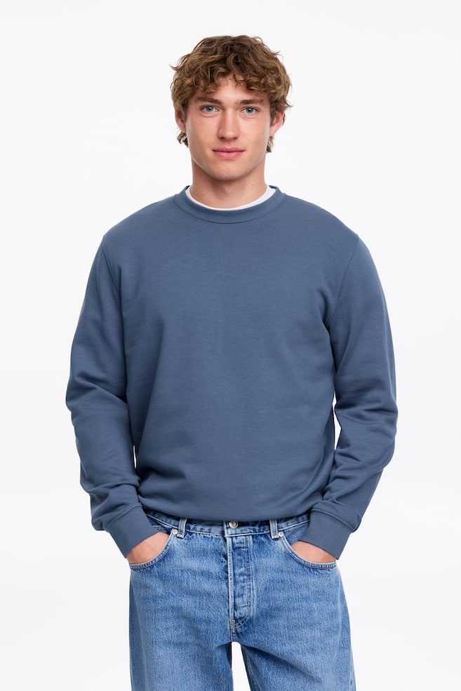 A slate blue crew neck sweatshirt is worn by a person facing forward. It features long sleeves with ribbed cuffs and is layered over a white t-shirt, visible at the neckline. - 1