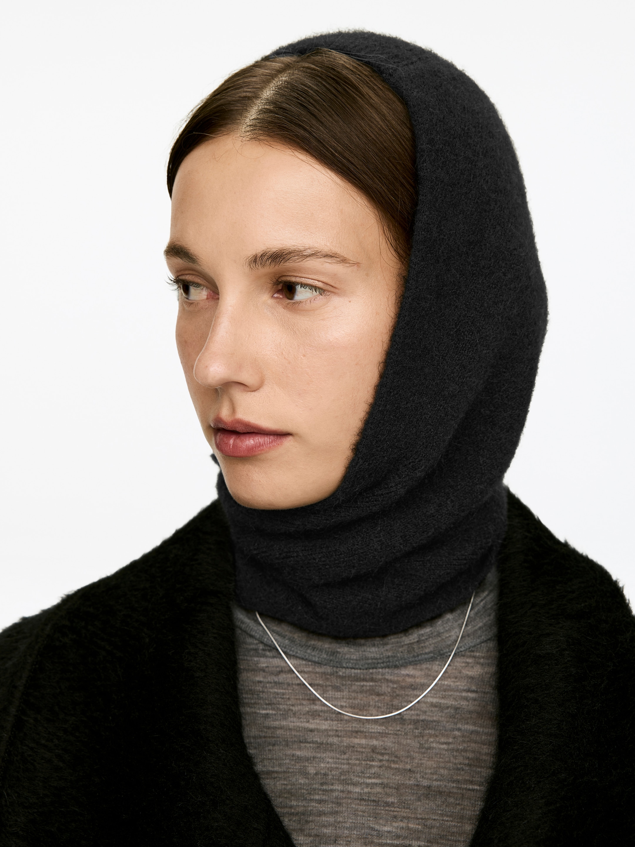 Ver imagen más grande: Una capucha de estilo pasamontañas de color gris oscuro, hecha de una mezcla de alpaca y lana, está siendo usada por el modelo. Está cubriendo la cabeza y el cuello del modelo, y se lleva sobre un jersey de cuello alto gris y debajo de una chaqueta oscura y texturizada.