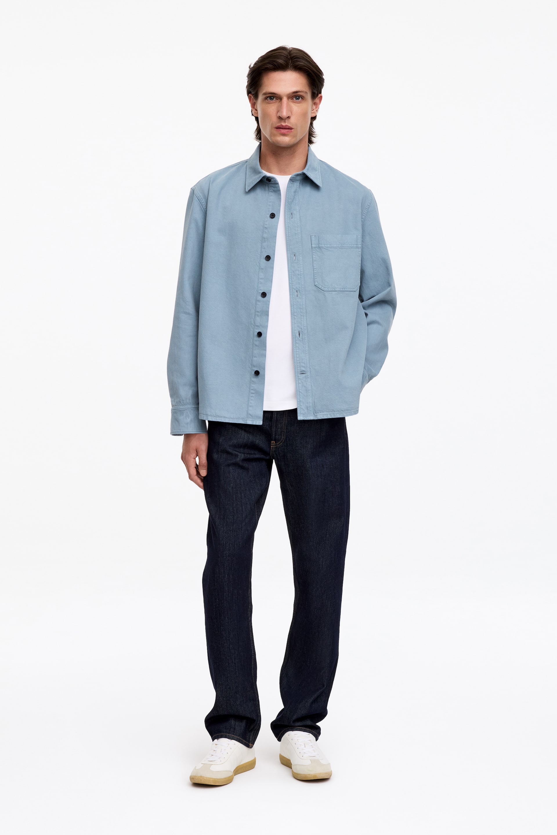 Visualizza immagine più grande: Il modello indossa una overshirt in twill azzurro chiaro lasciata sbottonata, caratterizzata da colletto classico, chiusura frontale con bottoni e tasca singola sul petto. È abbinata a una T-shirt bianca e a jeans blu scuro.