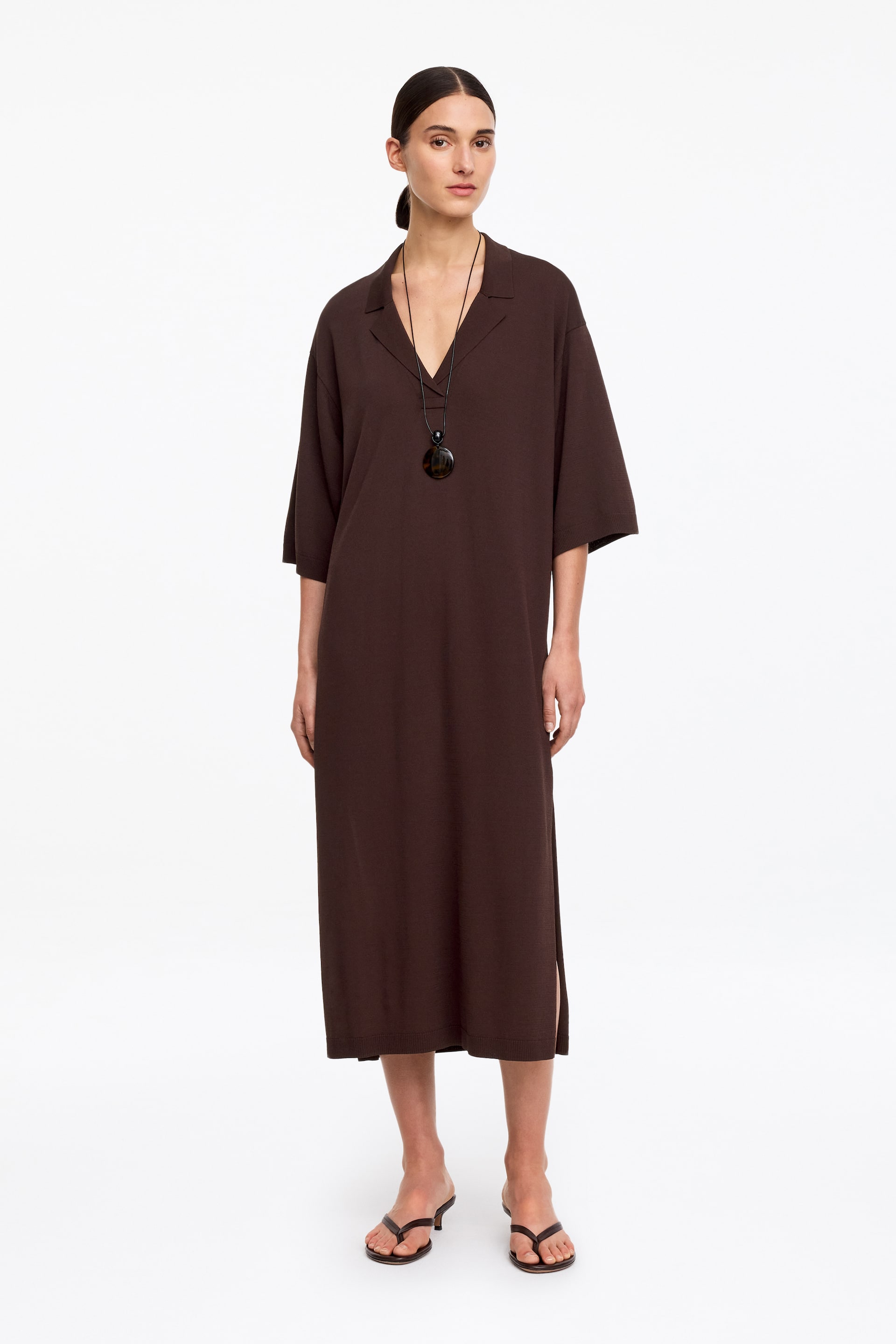 Agrandir l'image: La robe midi marron foncé présente un décolleté en V avec un col à revers, des manches trois-quarts larges et de petites fentes latérales, avec un ourlet fini en détail côtelé. Un collier pendentif rond et sombre orne le devant de cette robe à coupe ample.