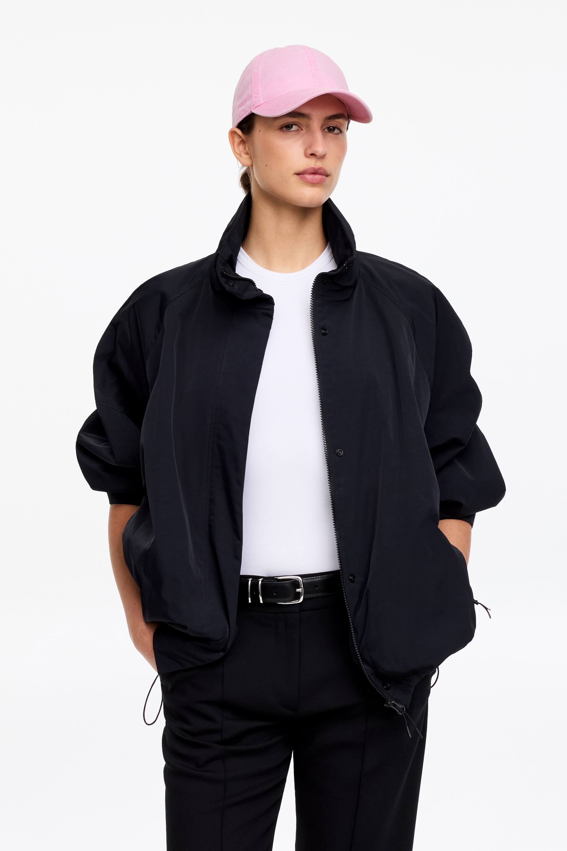 Agrandir l'image: Une veste noire présente une coupe décontractée avec des manches raglan volumineuses, un col montant et des poches latérales visibles. Elle est dotée d'une fermeture éclair frontale complète dissimulée par des boutons-pression, et un cordon de serrage est visible à l'ourlet, car elle est portée ouverte sur un t-shirt blanc.