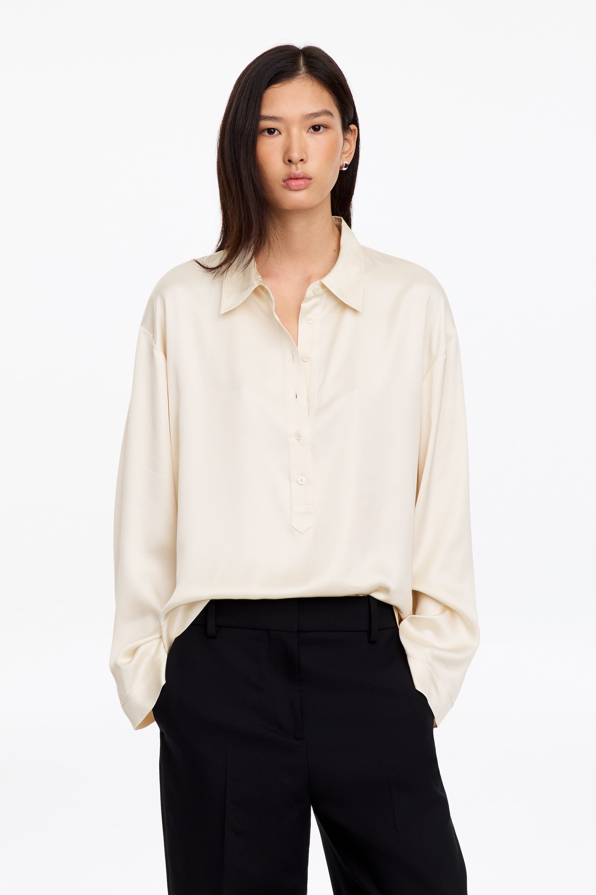 Se større billede: En råhvid poloshirt med lange ærmer, afslappet pasform, sænkede skuldre, klassisk krave og halv stolpelukning med knapper bæres af modellen. Skjorten er stylet løst nedstukket i sorte bukser, hvilket fremhæver dens bløde og flydende fald.