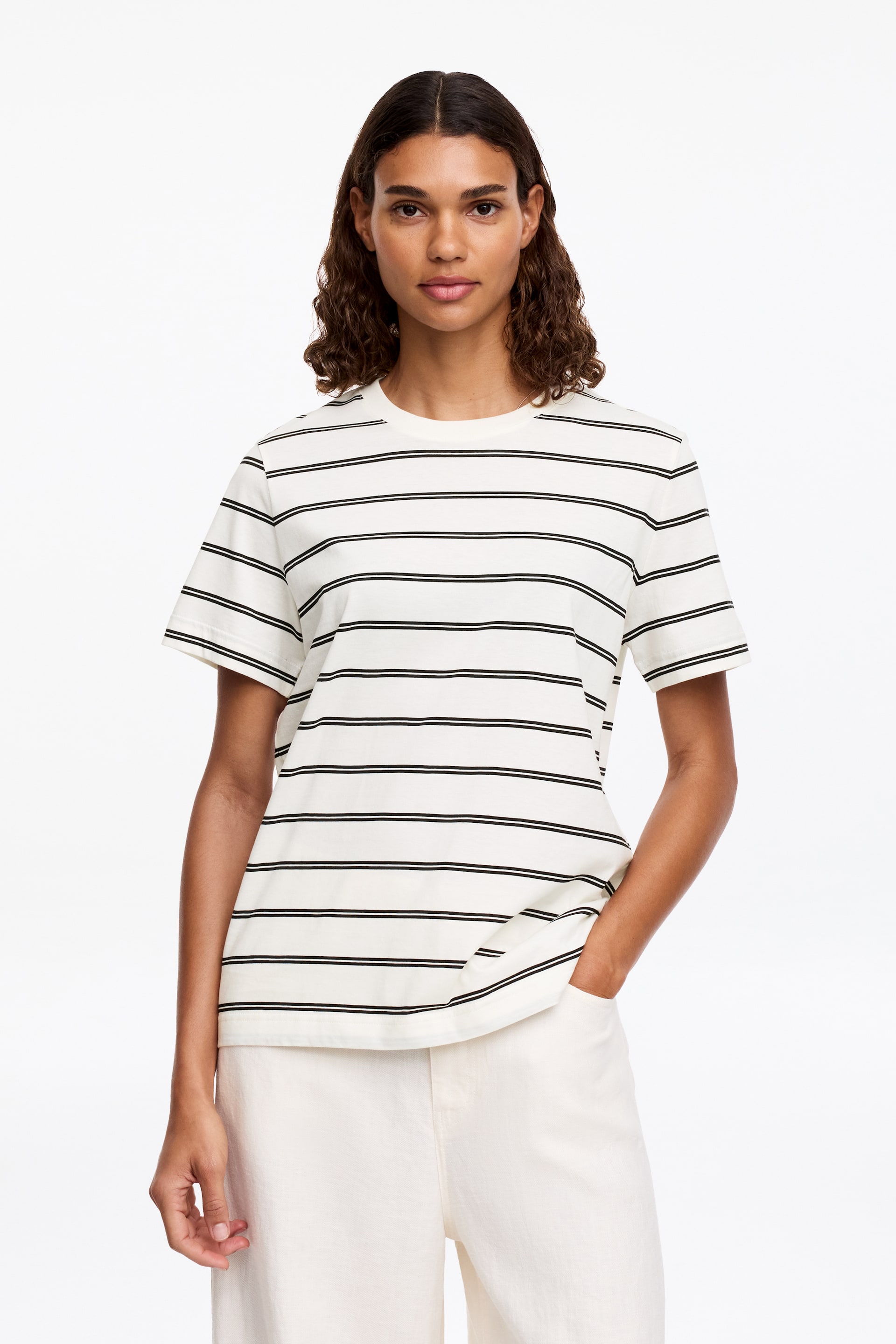 Visa större bild: En off-white t-shirt med tunna svarta horisontella ränder bärs av modellen. T-shirten har en ribbad rund halsringning och korta ärmar, och faller över ljusa byxor. Modellens vänstra hand vilar i en byxficka.