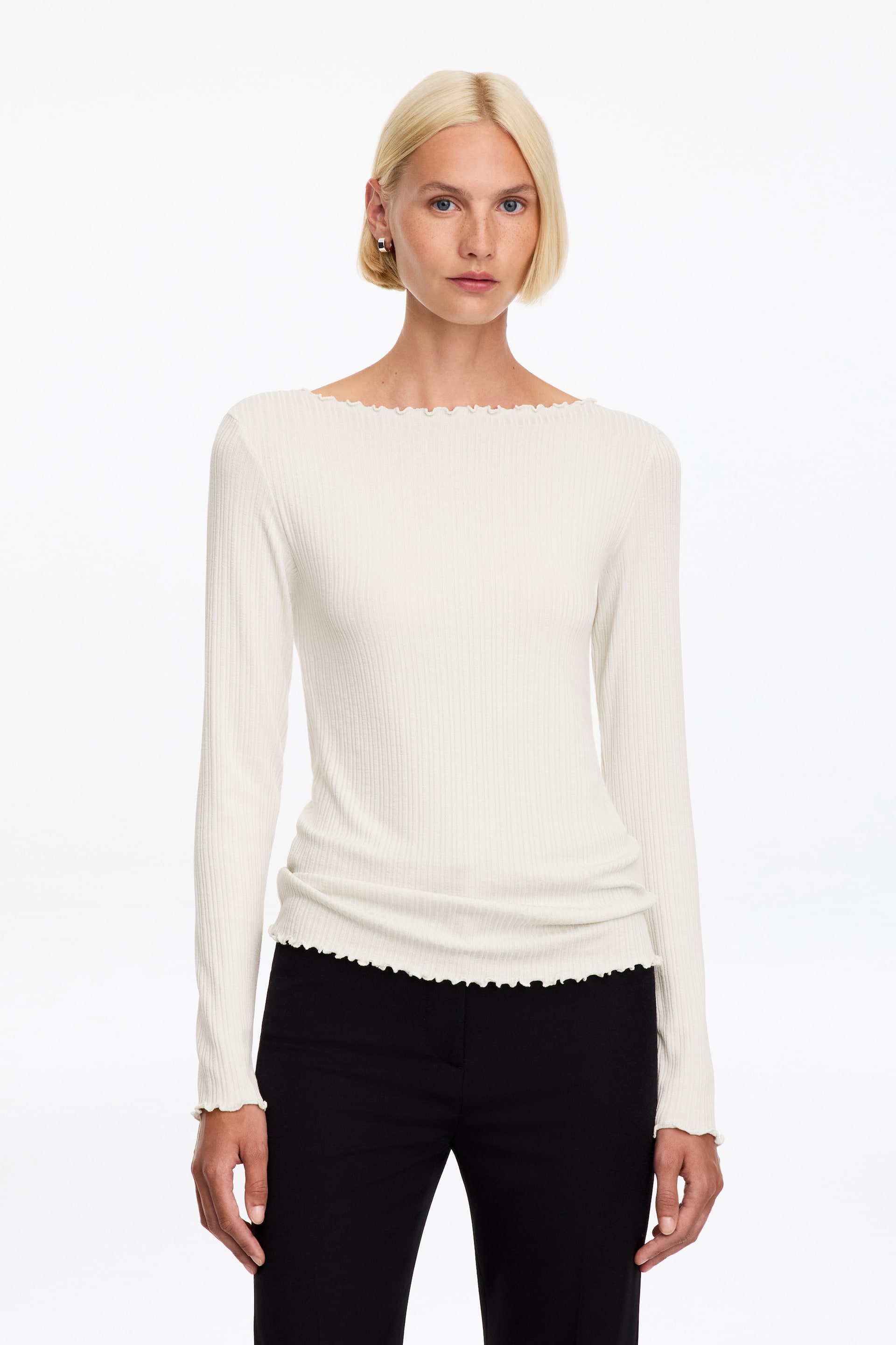 Visualizza immagine più grande: Una maglia a maniche lunghe color bianco sporco, in maglia a coste, indossata dalla modella, con vestibilità aderente e scollo a barchetta rifinito con volant babylock su scollo, polsini e orlo. La maglia è abbinata a pantaloni neri.