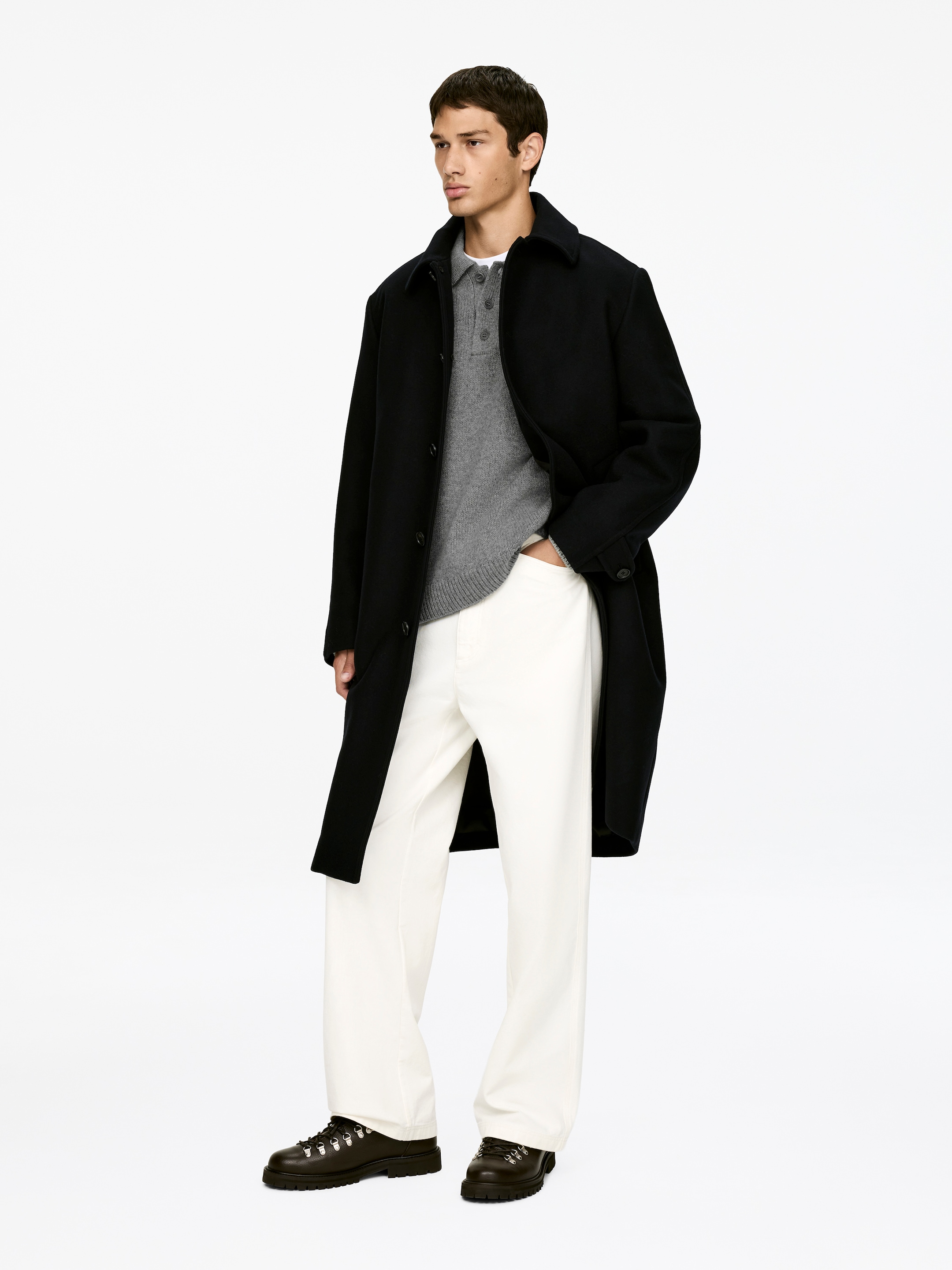 Agrandir l'image: Un manteau noir à simple boutonnage avec un col classique est présenté, superposé à un pull polo gris à manches longues et un t-shirt à col rond blanc. Cette tenue est complétée par un pantalon crème à jambes larges et des chaussures à lacets sombres et robustes avec des œillets en D, tandis que le mannequin est légèrement incliné vers la droite et regarde à droite.