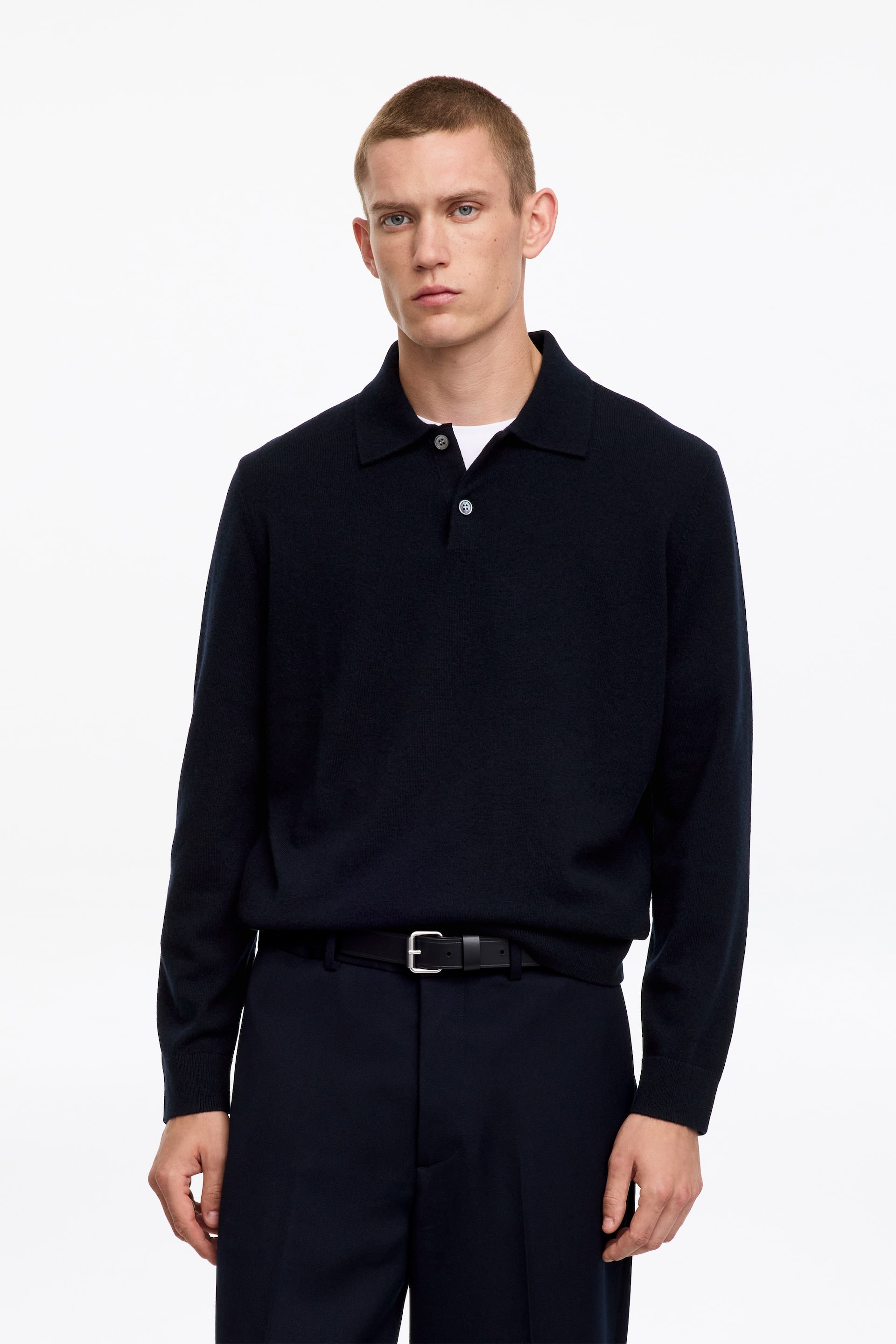 View larger image: A navy fine-knit polo shirt with a classic collar and a buttoned half placket is being worn by the model. It features mother-of-pearl buttons, is layered over a white undershirt, and is tucked into dark trousers.