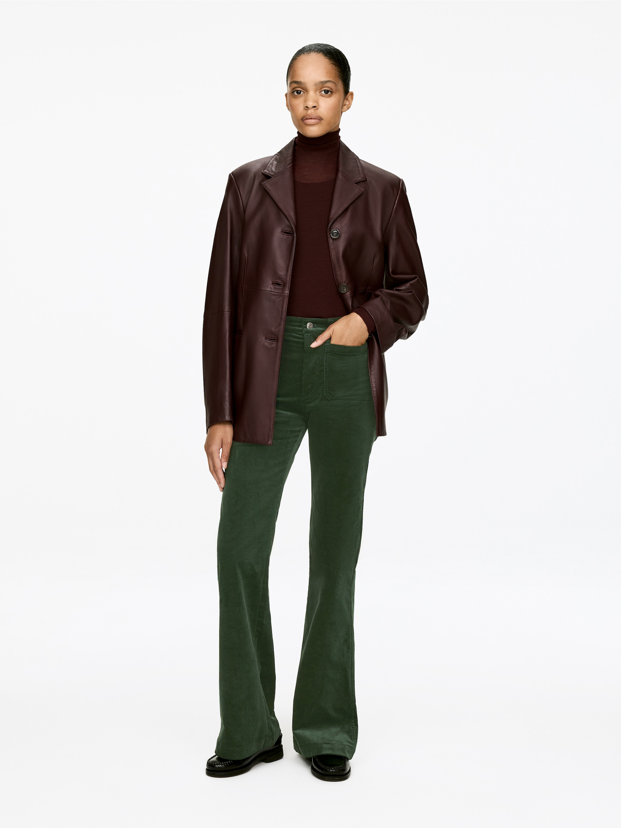 Ver imagen más grande: Un blazer de cuero marrón oscuro se presenta de frente, llevado abierto sobre un suéter de cuello alto rojo oscuro y pantalones de pana de campana verde oscuro. El blazer monocruzado presenta solapas en punta y tres botones frontales, y los pantalones muestran bolsillos utilitarios frontales y una apertura de pierna ancha.