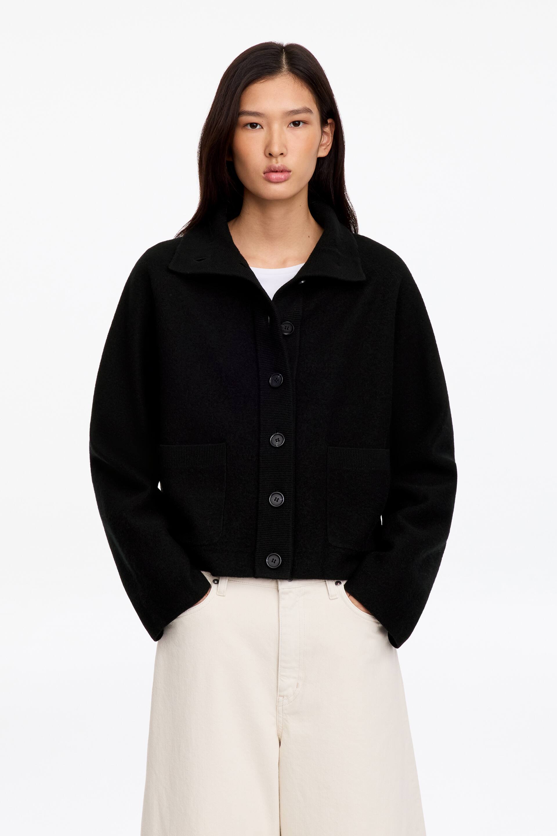 Agrandir l'image: Un cardigan en laine bouillie noir est porté par le mannequin, avec une patte de boutonnage, un col à pointes, des manches raglan et des poches plaquées sur le devant. Le cardigan est porté sur un t-shirt blanc et associé à un pantalon blanc cassé.