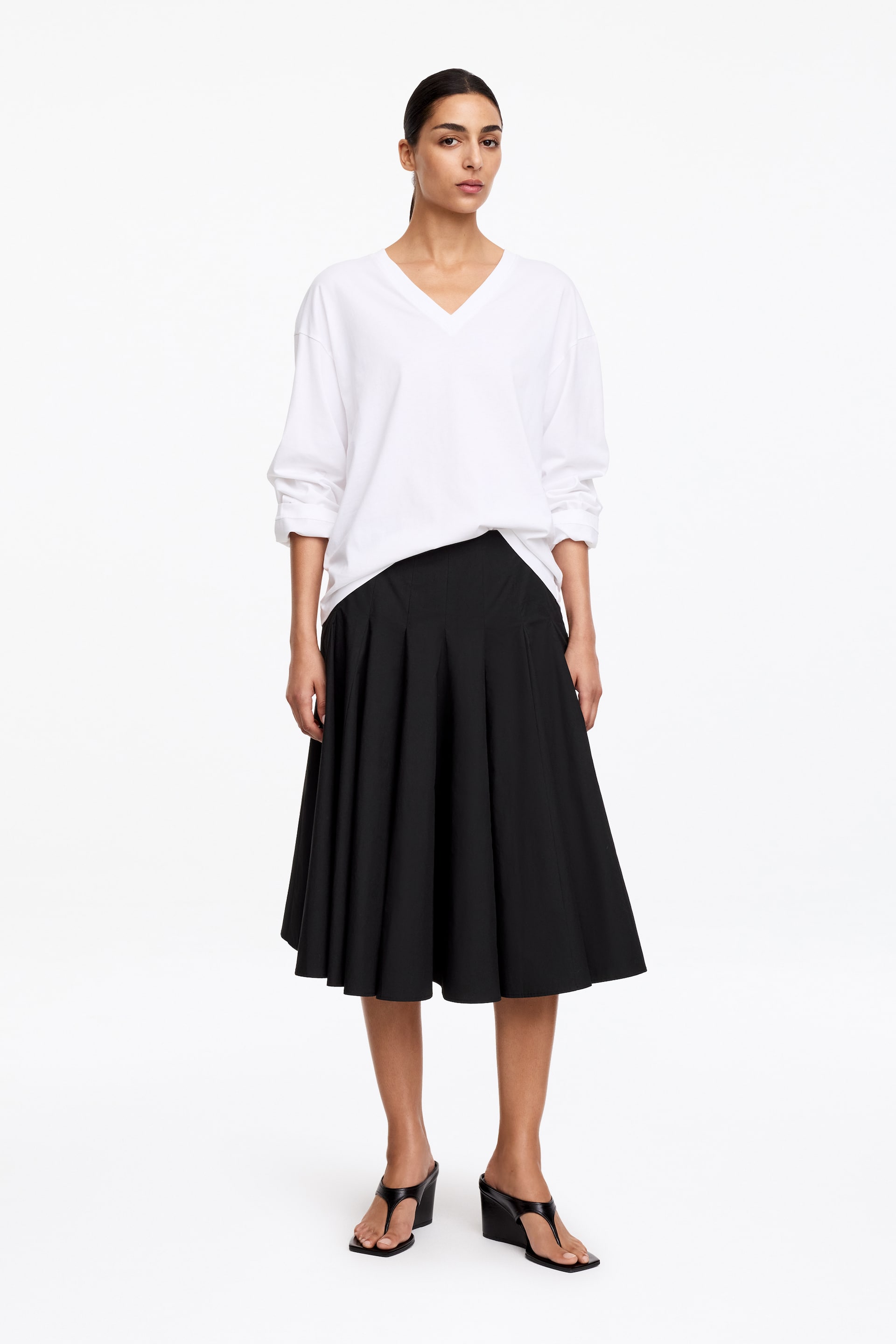 Agrandir l'image: Une jupe midi noire est présentée, dotée d'une taille haute et d'une silhouette évasée en ligne A. La taille de la jupe est partiellement masquée par un haut blanc à manches longues rentré à l'avant.