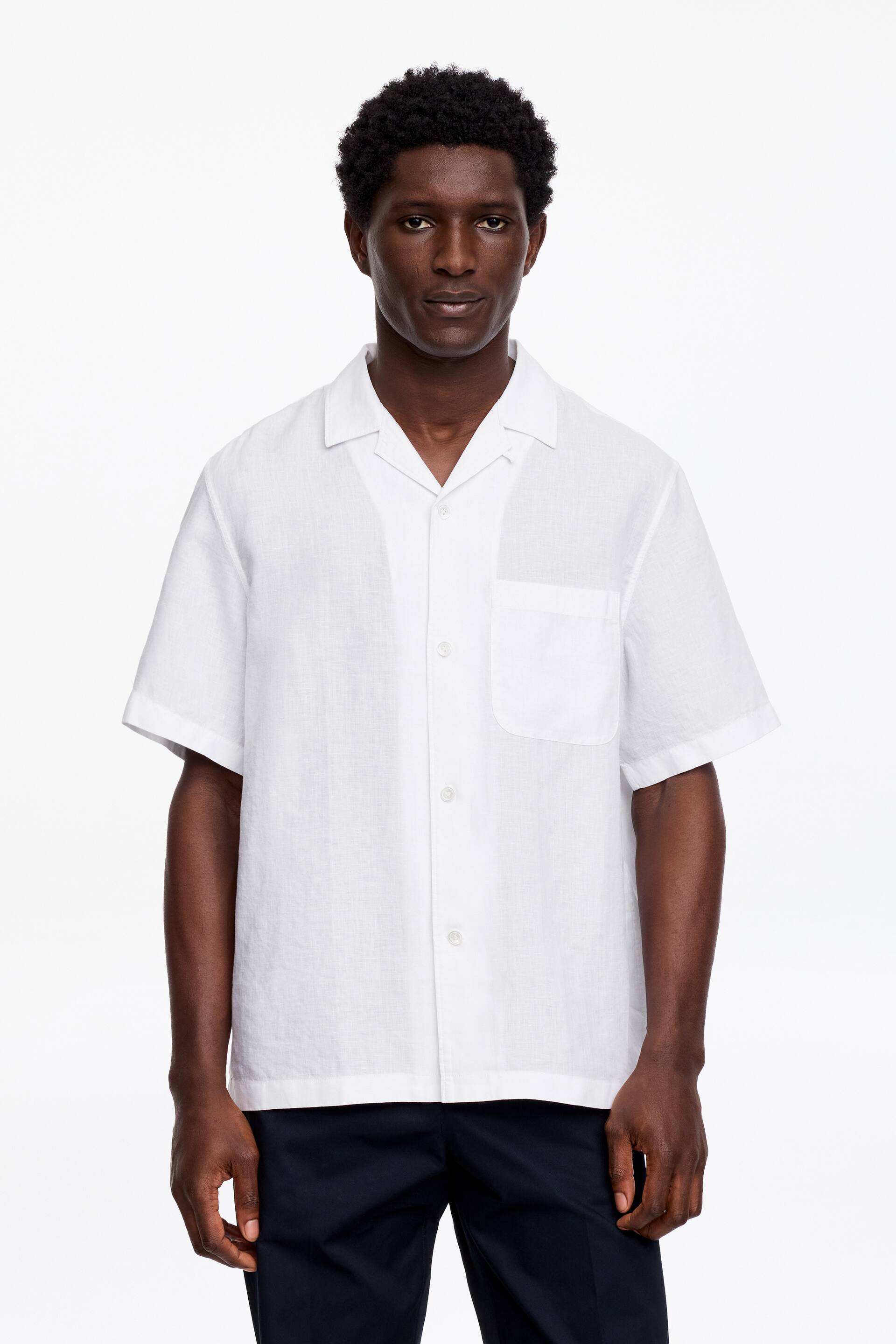 Se større billede: En hvid kortærmet linnedskjorte bæres af modellen. Den har en camp-krave, knaplukning foran og en brystlomme på venstre side.