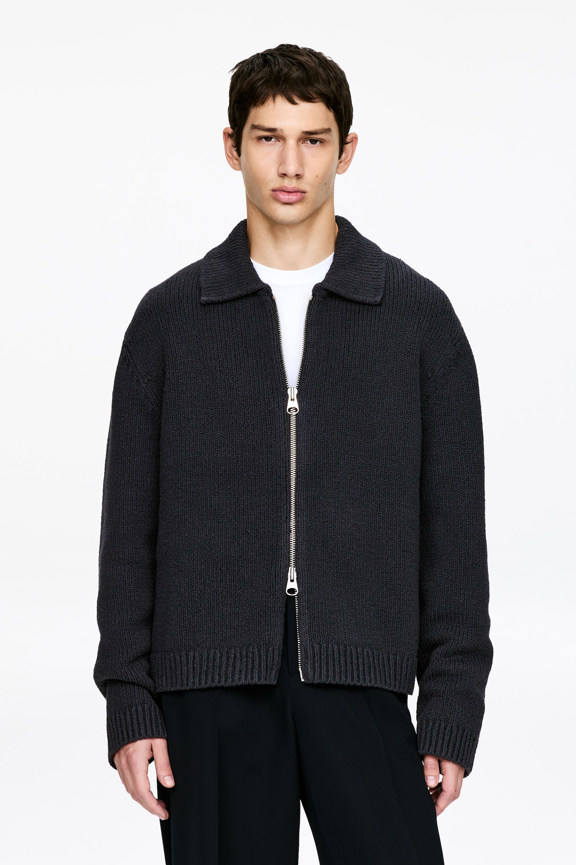 Visualizza immagine più grande: Una giacca a maglia grigio scuro con zip è presentata frontalmente. Presenta un colletto aperto, una cerniera a tutta lunghezza color argento e polsini e orlo a coste, ed è indossata sopra una t-shirt bianca con scollo a girocollo e pantaloni neri.