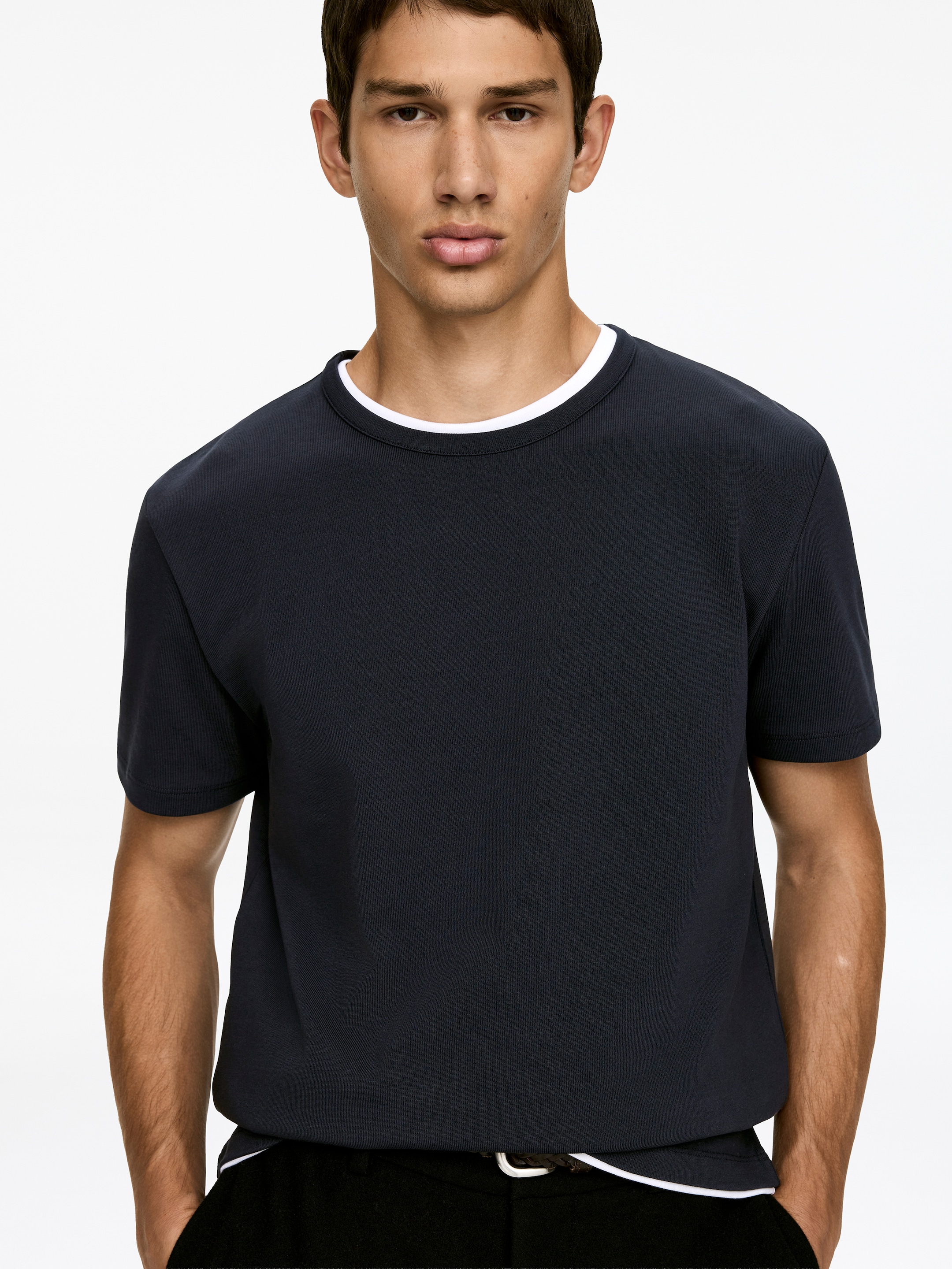 Visa större bild: En mörkblå T-shirt med rund hals, tillverkad av bomull, bärs av modellen. Den har en åtsittande passform vid bröstet och armarna, och ett vitt plagg är lagrad under dess halsringning och fåll.