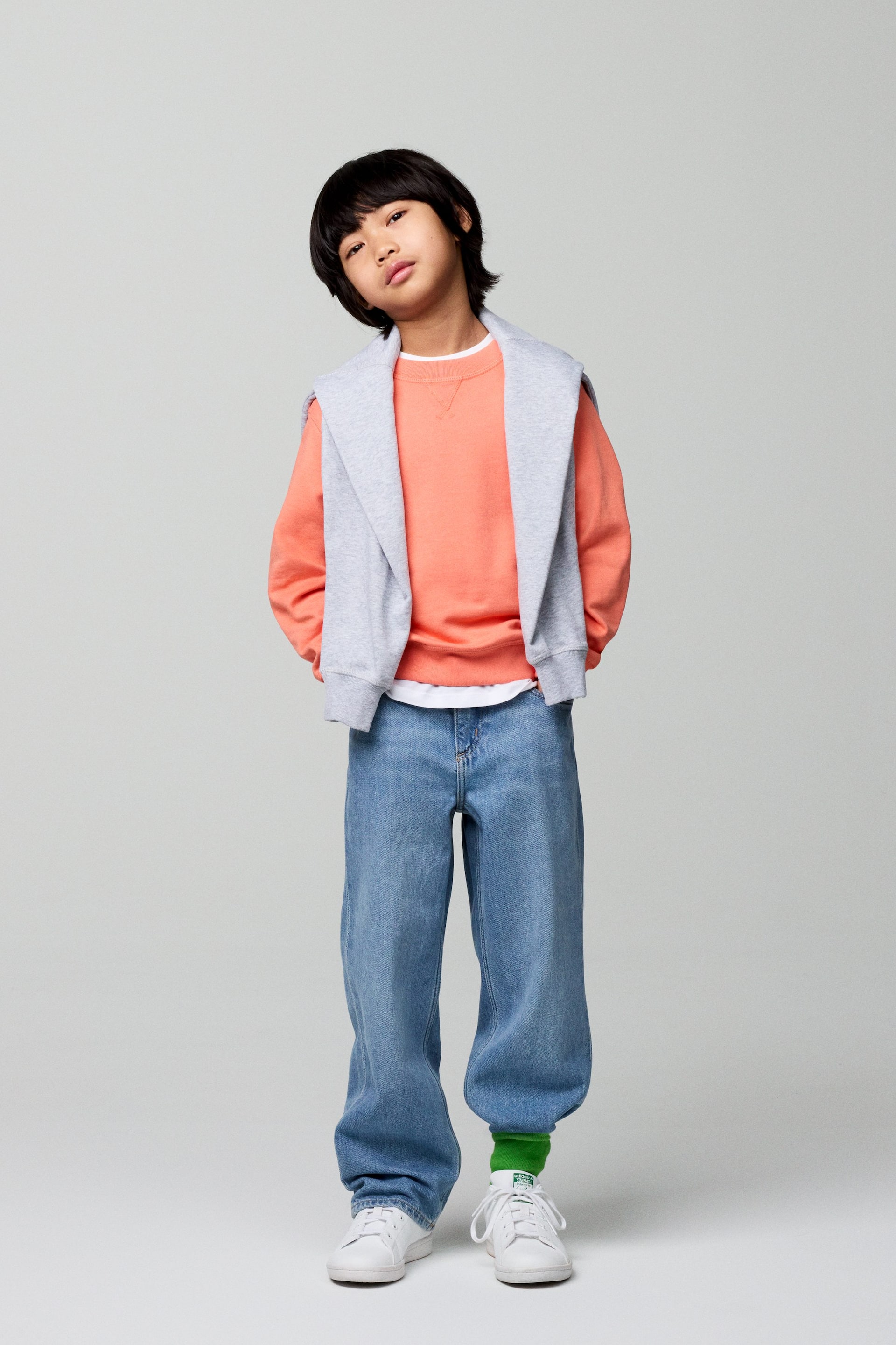 Grotere afbeelding bekijken: Een oranje crewneck sweatshirt met geribde boorden en zoom, gedragen over een wit t-shirt, met een grijze capuchontrui nonchalant over de schouders gedrapeerd. Loszittende lichtblauwe jeans bedekken de benen, waarbij felgroene sokken en witte vetersneakers zichtbaar zijn.